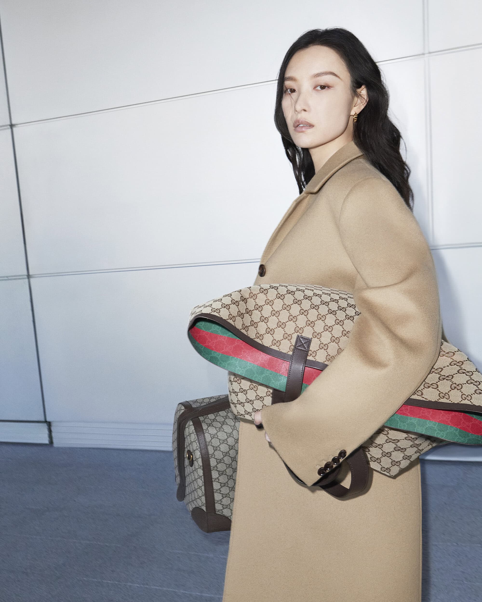 Elegant Asian woman in a beige coat carrying Gucci monogrammed bags, showcasing fashion and style against a minimalist white background.