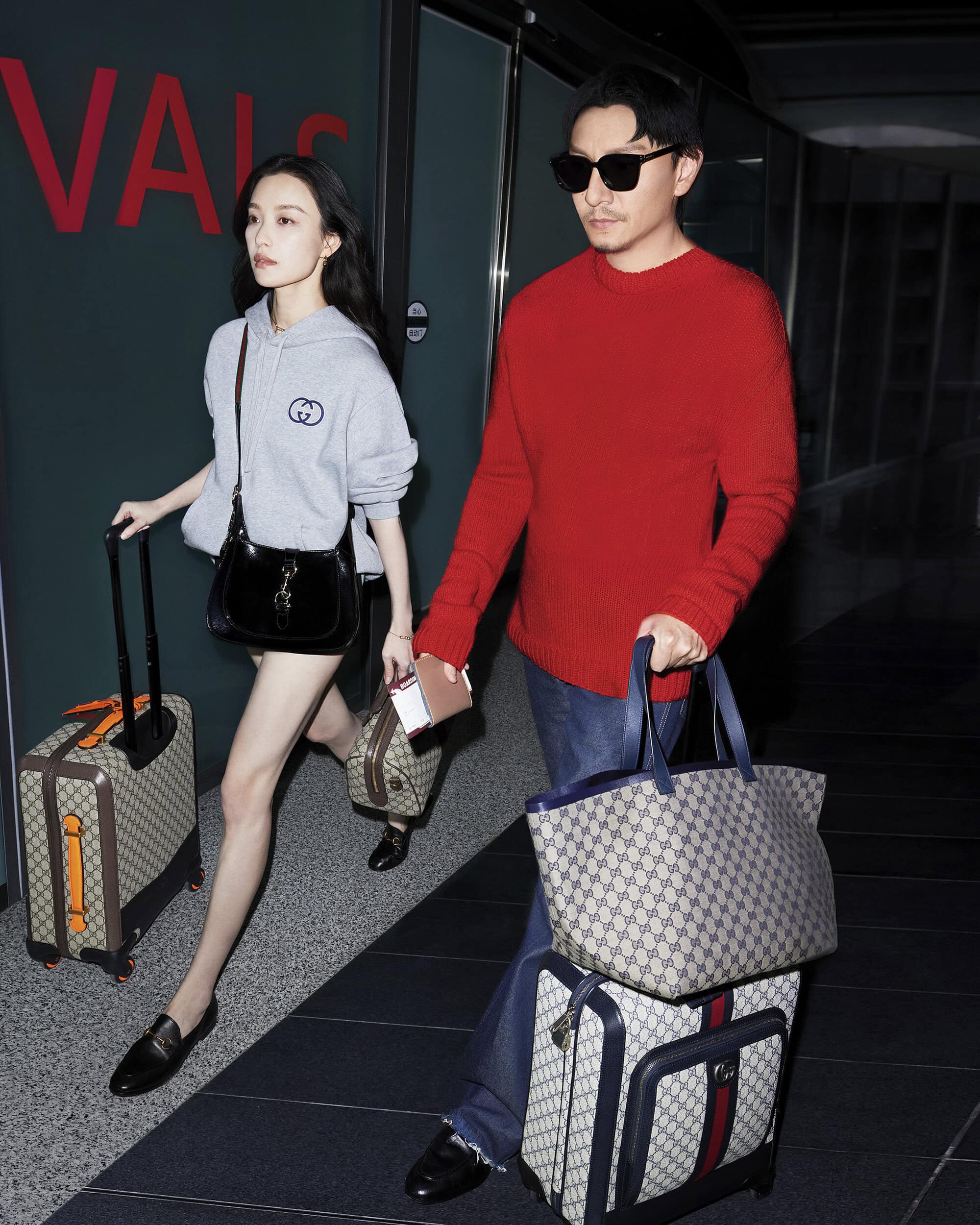 A couple walks through an airport, pulling matching luggage, wearing casual clothing. The woman wears a grey hoodie, the man a red sweater.