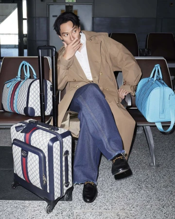 Adult male in a trench coat sits next to designer luggage in a modern airport departure lounge with neutral colors and minimalist design.