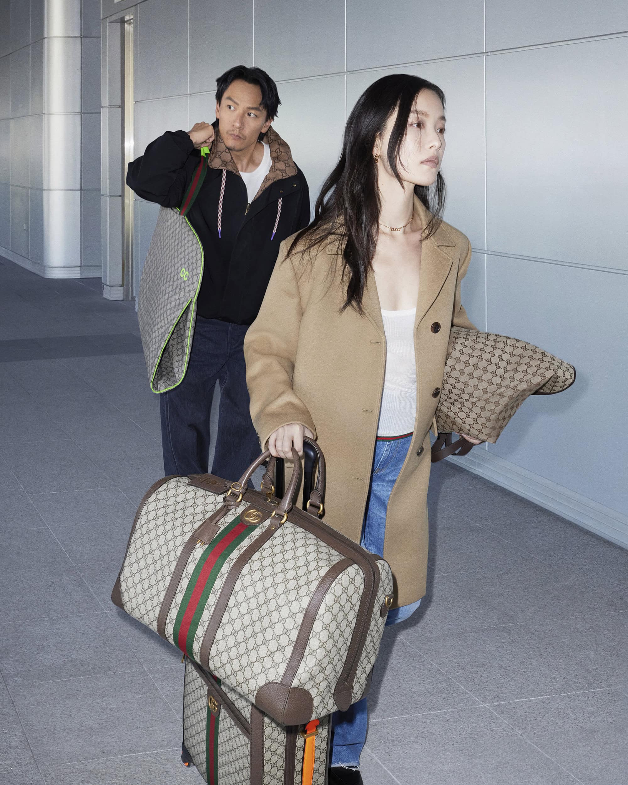 Two people in transit carrying patterned travel bags and a suitcase, walking through a modern airport or train station with neutral colors.