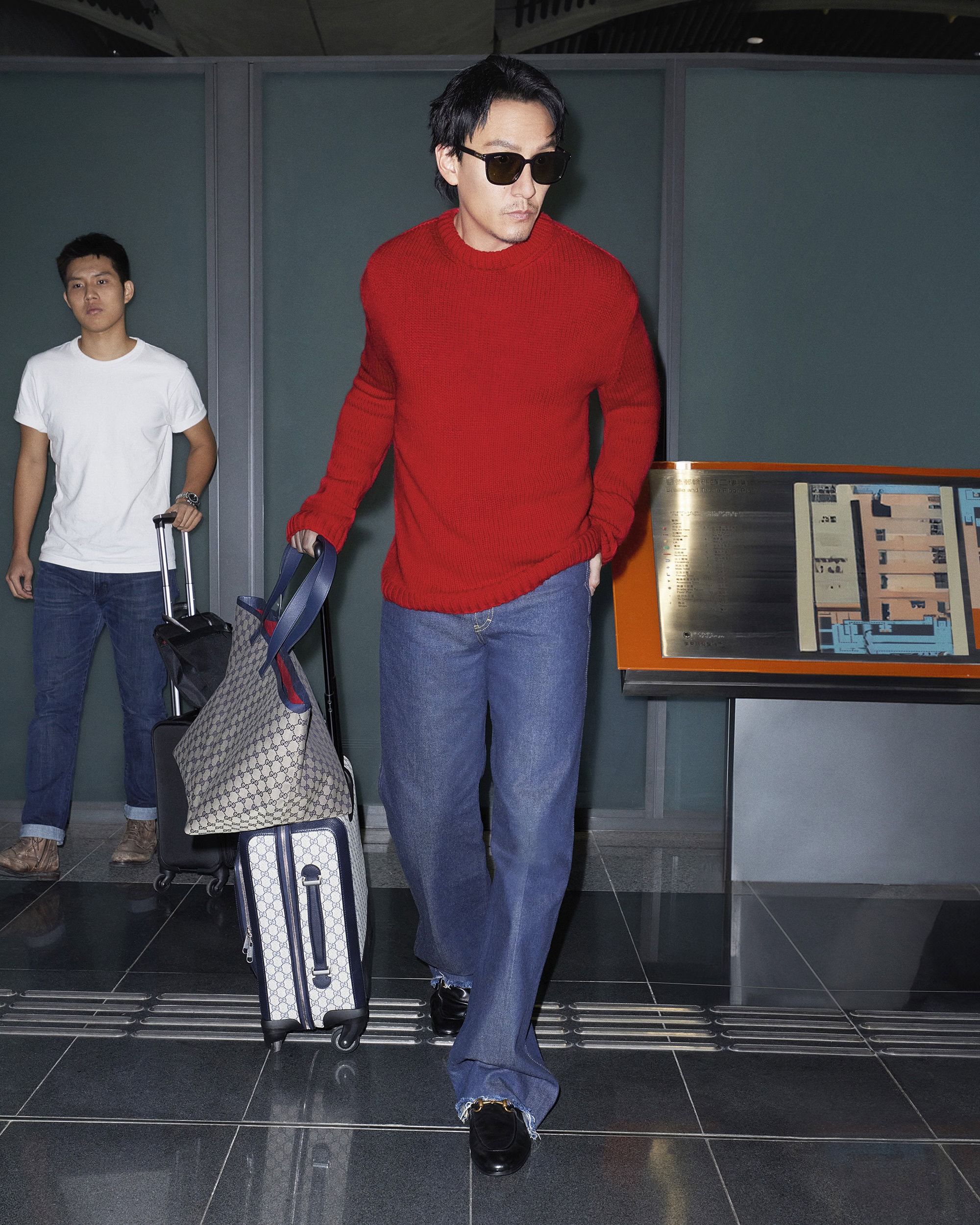 Adult male wearing sunglasses and a red sweater walks indoors with luggage. Another man is in the background also pulling luggage.