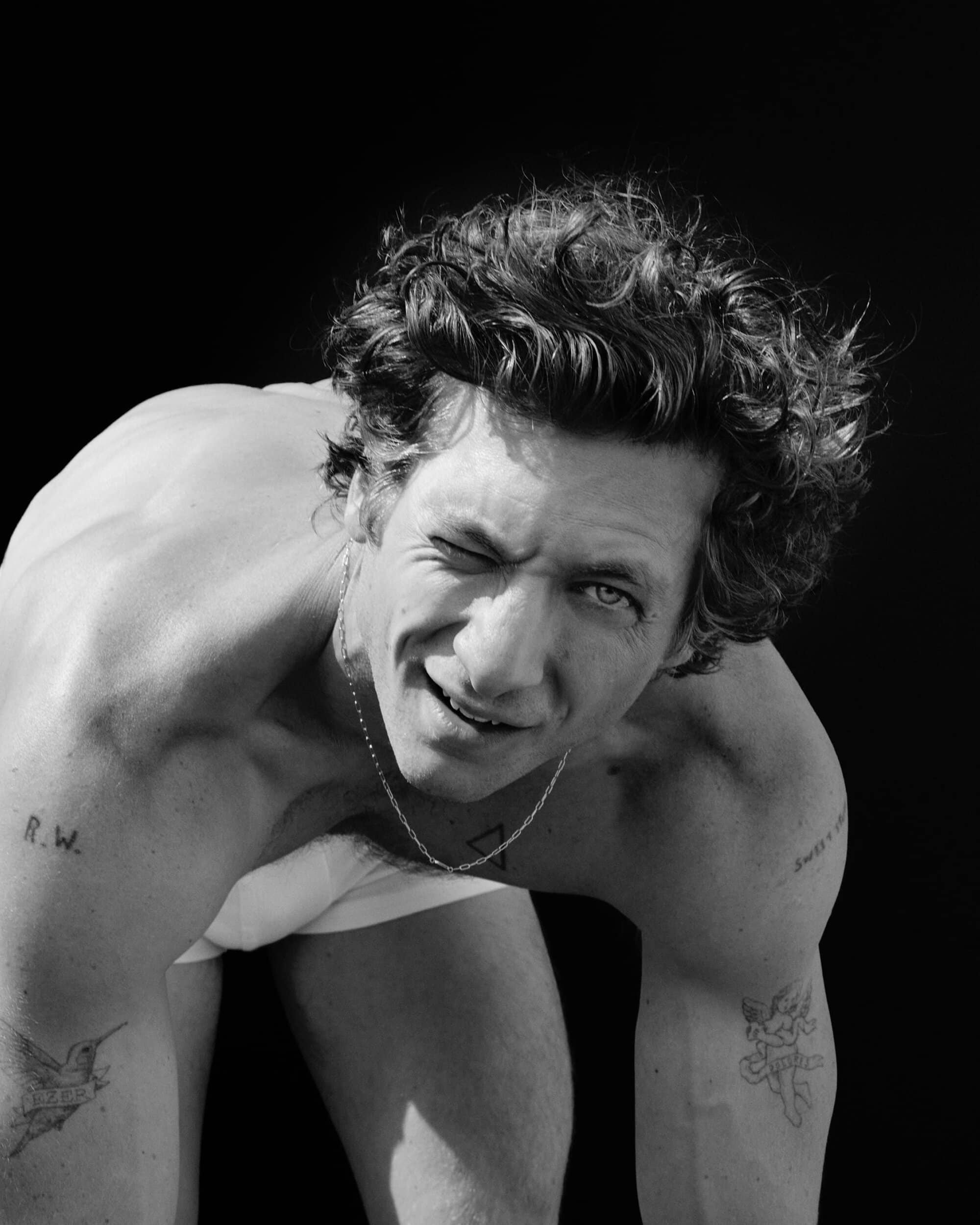Monochrome portrait of adult male winking at the camera. He has curly hair and tattoos on his arms and chest, against a black background.