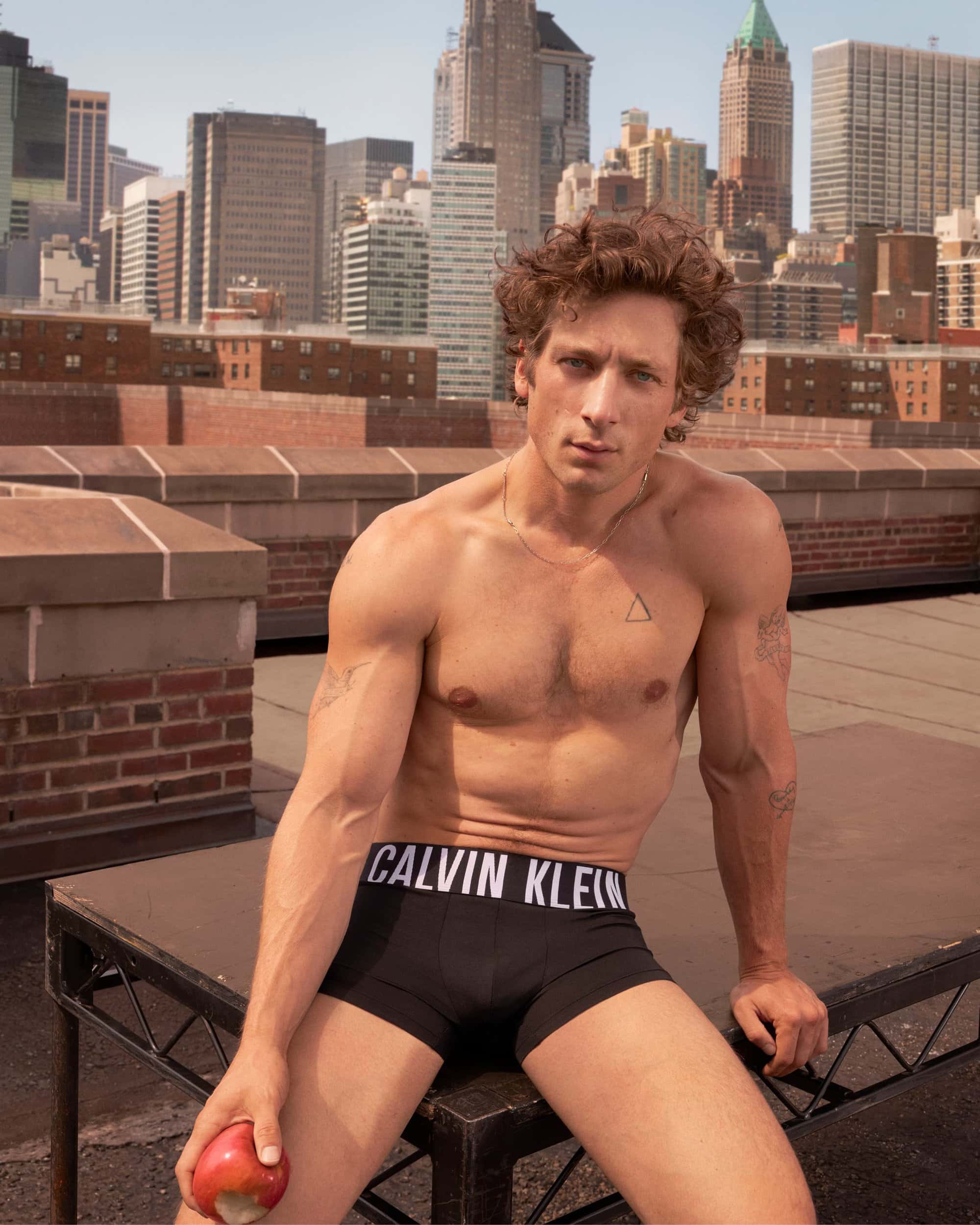 Shirtless man with Calvin Klein underwear sits on rooftop in a cityscape, holding an apple, under natural light, portrait orientation.