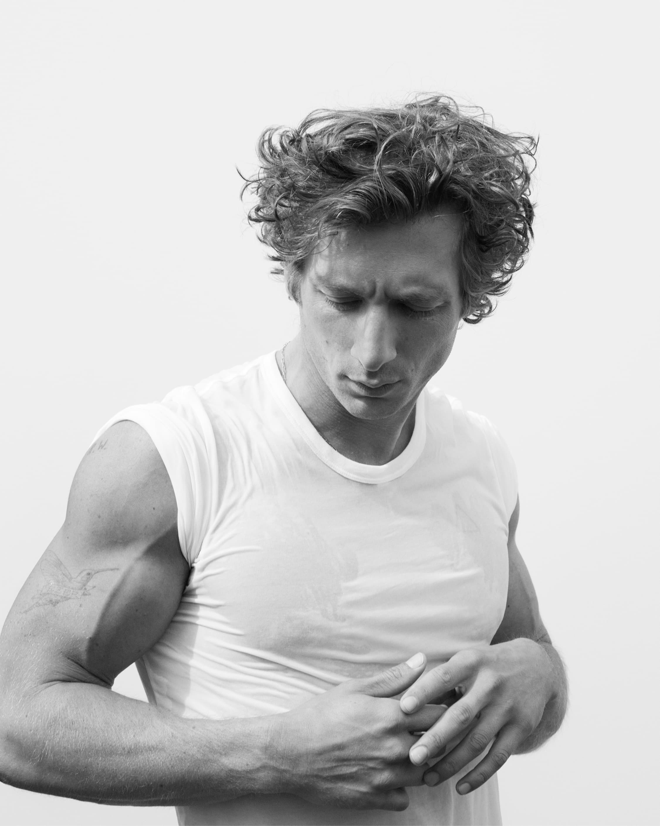 Monochrome portrait of an adult male with curly hair wearing a wet t-shirt, looking down with arms crossed in a bright, neutral setting.