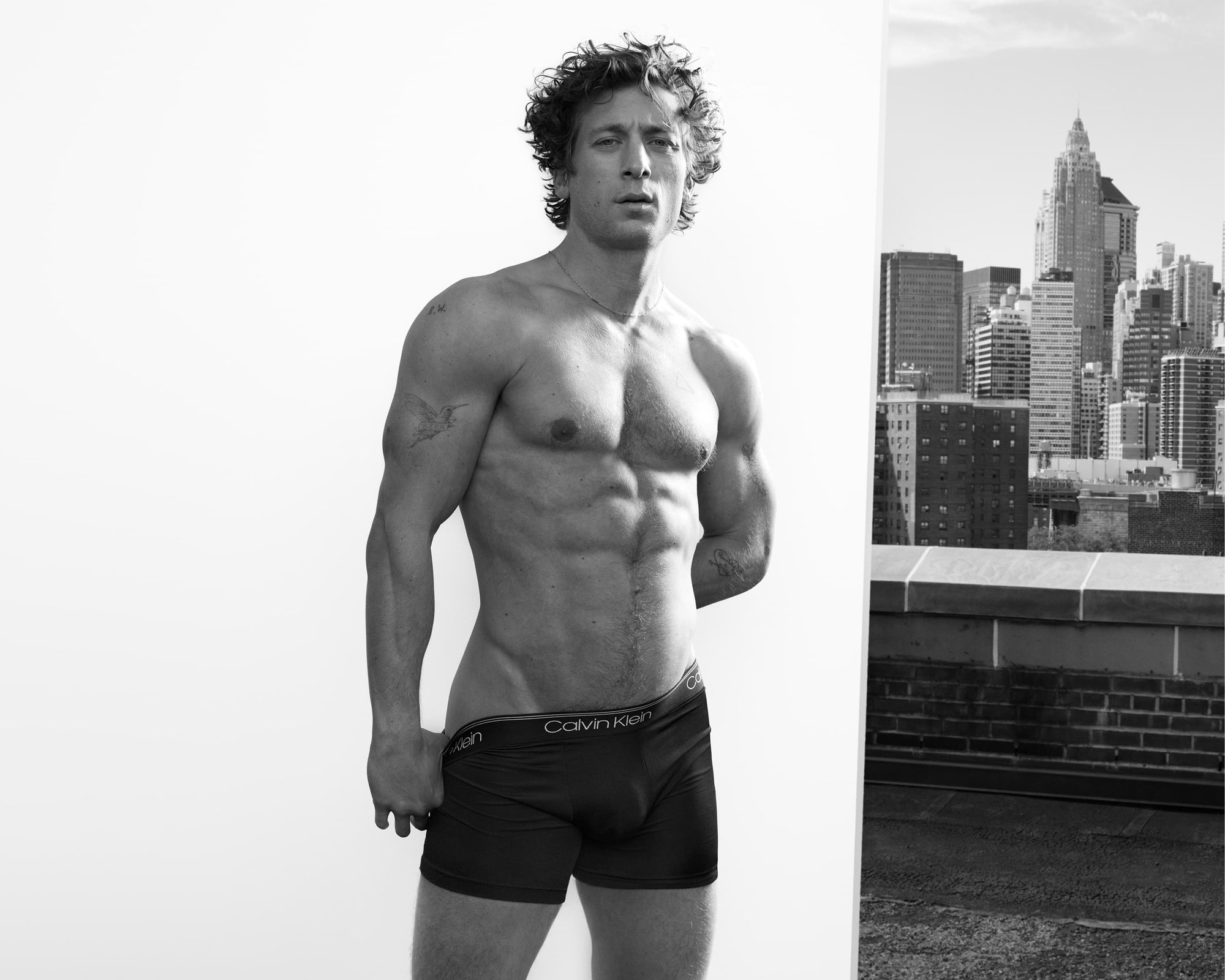 Black and white portrait of a muscular adult male posing in Calvin Klein underwear against a backdrop of urban cityscape buildings.