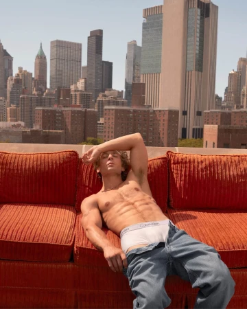 Man lying shirtless on an orange couch outdoors with a city skyline backdrop on a sunny day, posing with jeans and white underwear.