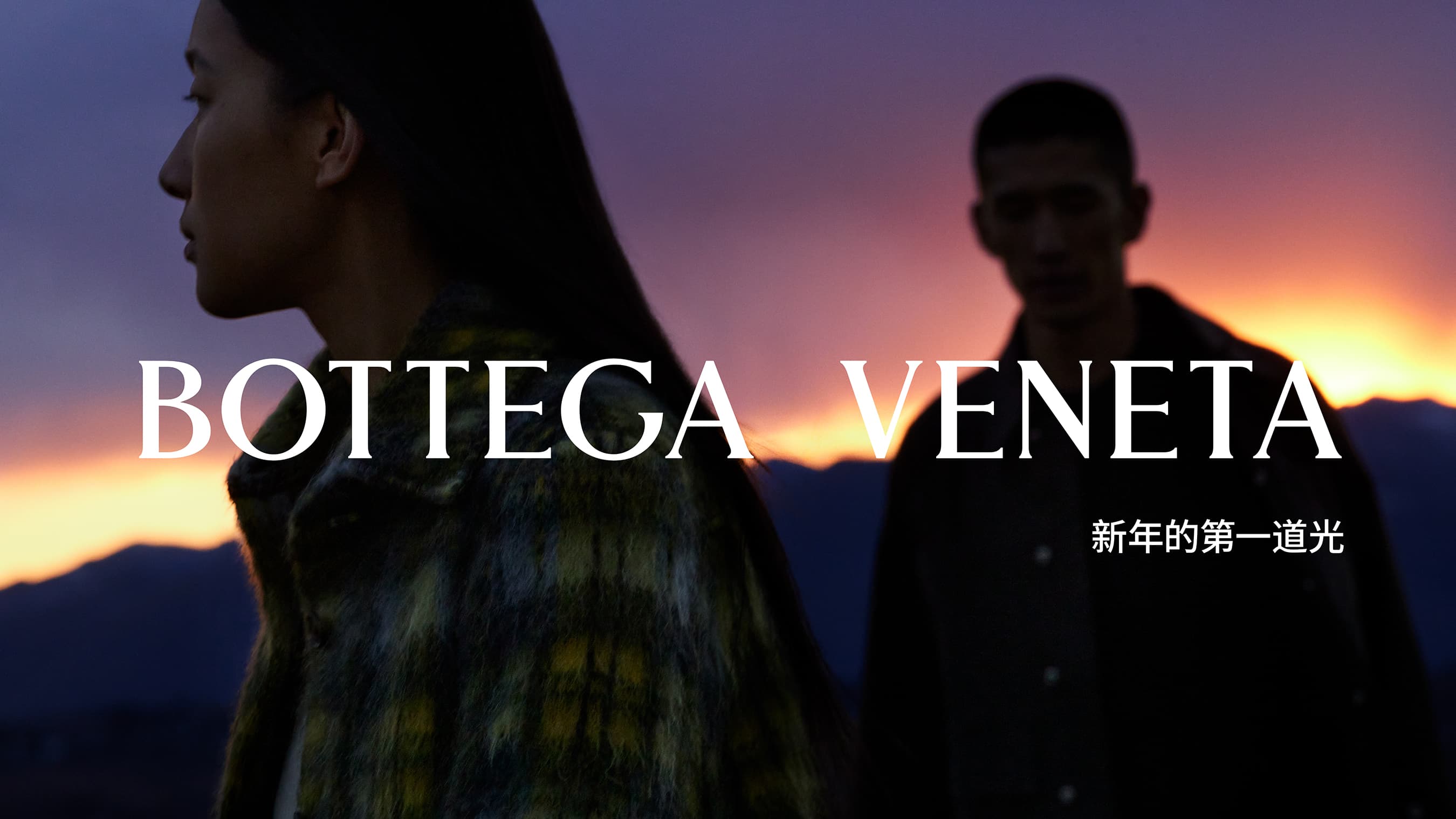 Fashion shot of a couple in Bottega Veneta clothing at dusk, standing outside with mountains in the background. Evening sky is visible.