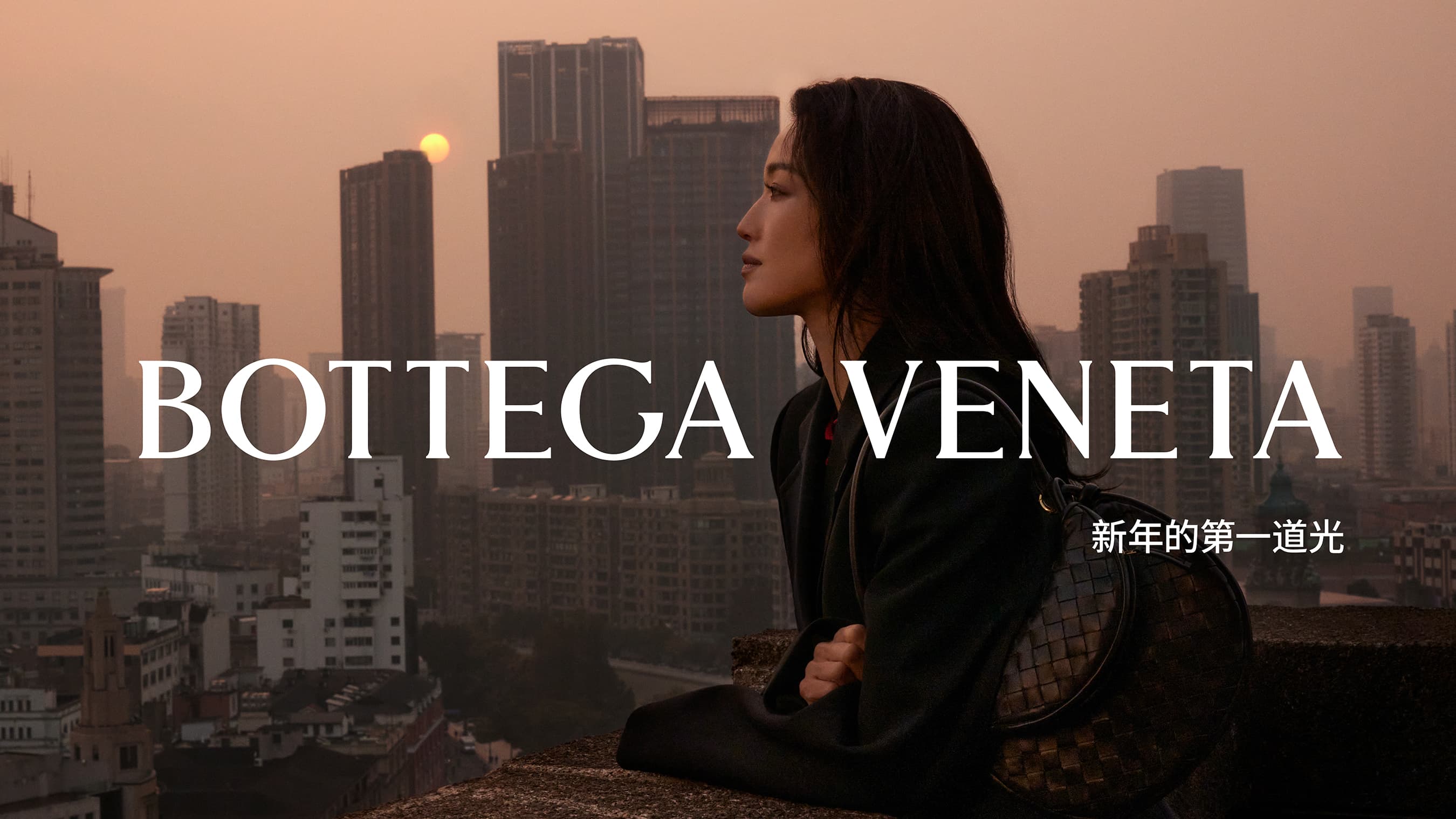 Asian woman in Bottega Veneta fashion, seated with purse, city skyline backdrop at sunset, elegant and stylish urban setting.