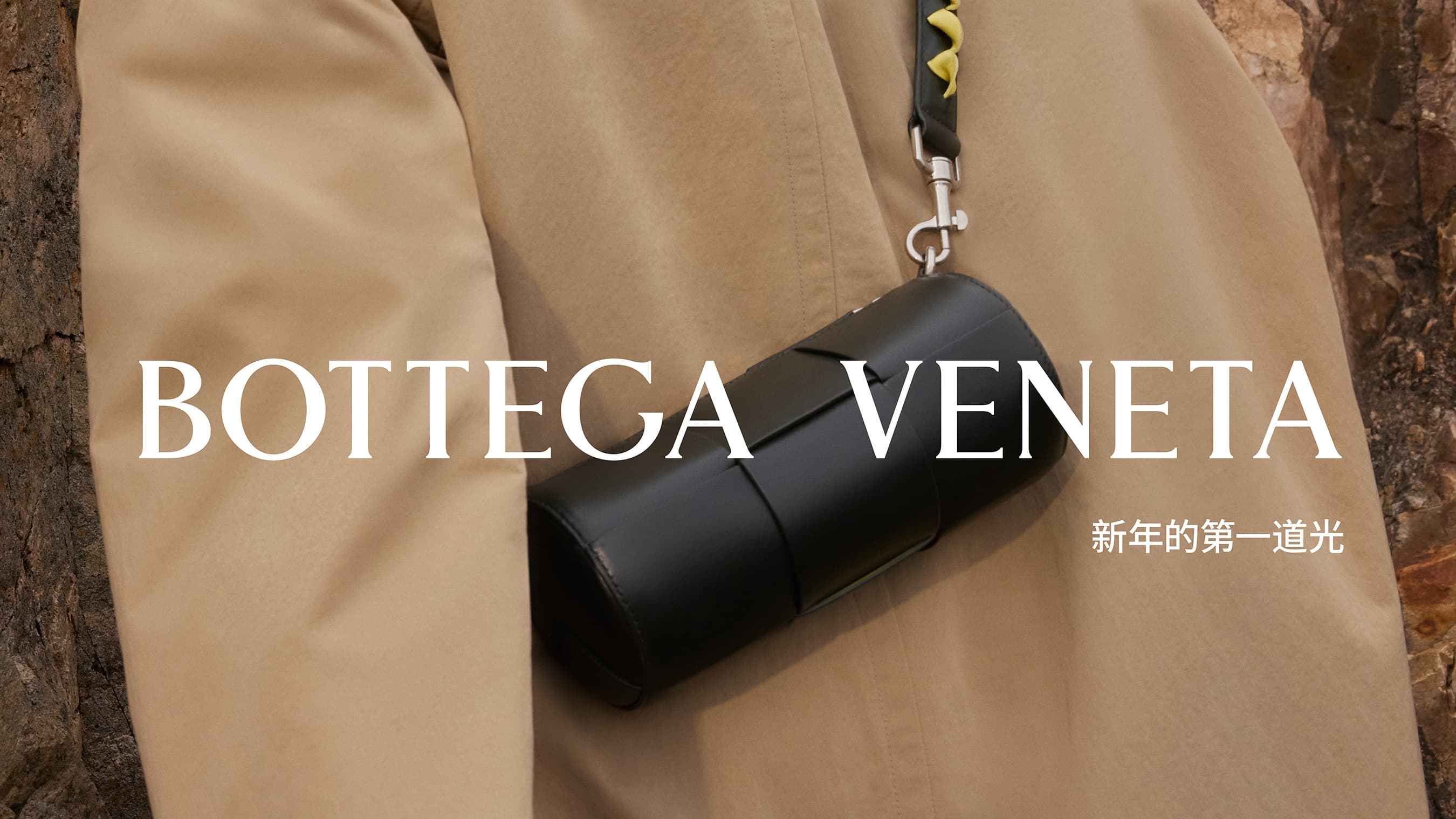 Black Bottega Veneta cylinder purse on shoulder strap, tan jacket, and blurred brown-toned woodgrain background in an ad.