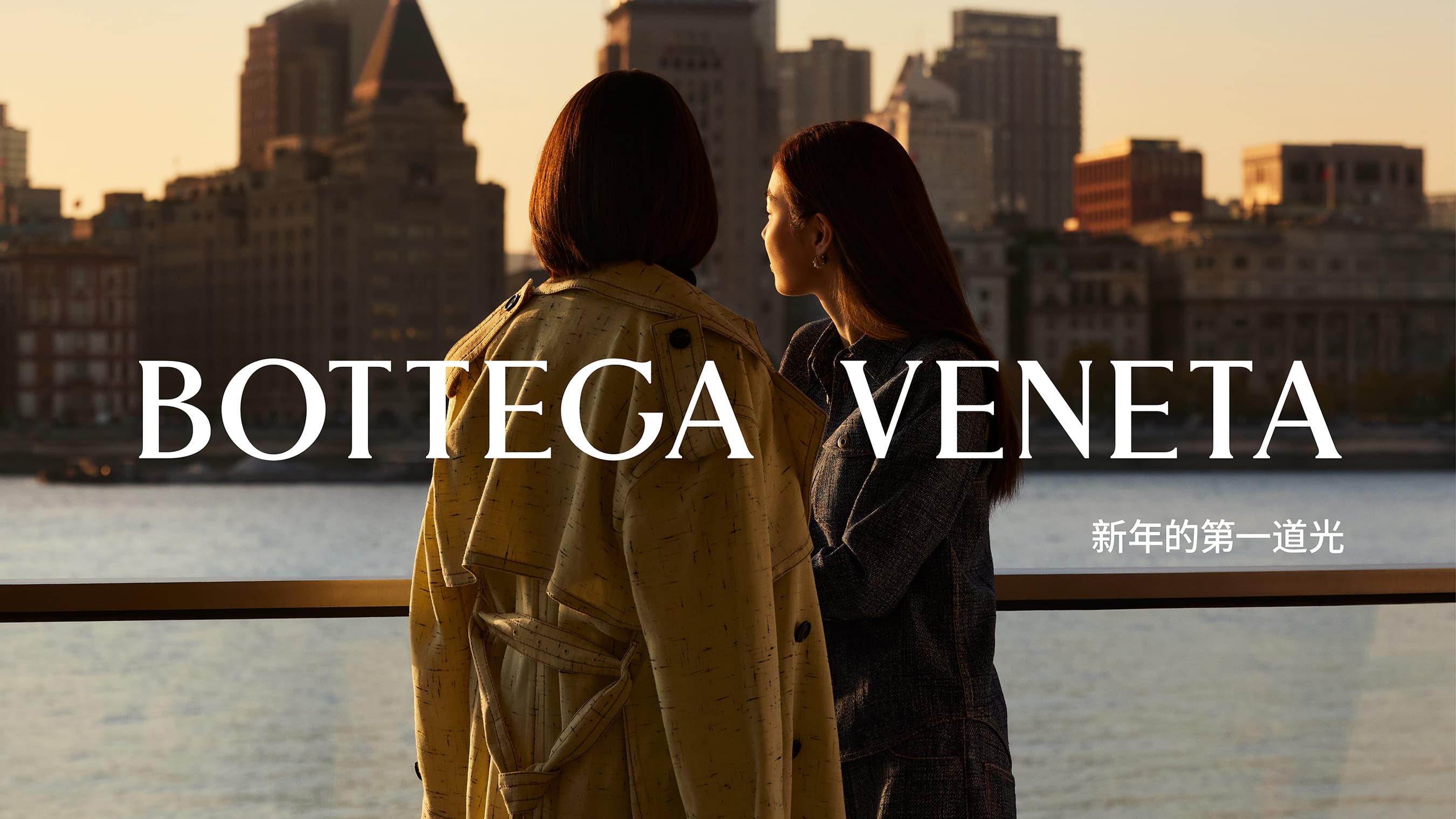 Two women in Bottega Veneta coats stand near a railing overlooking an urban skyline at sunset, captured in a fashion editorial.