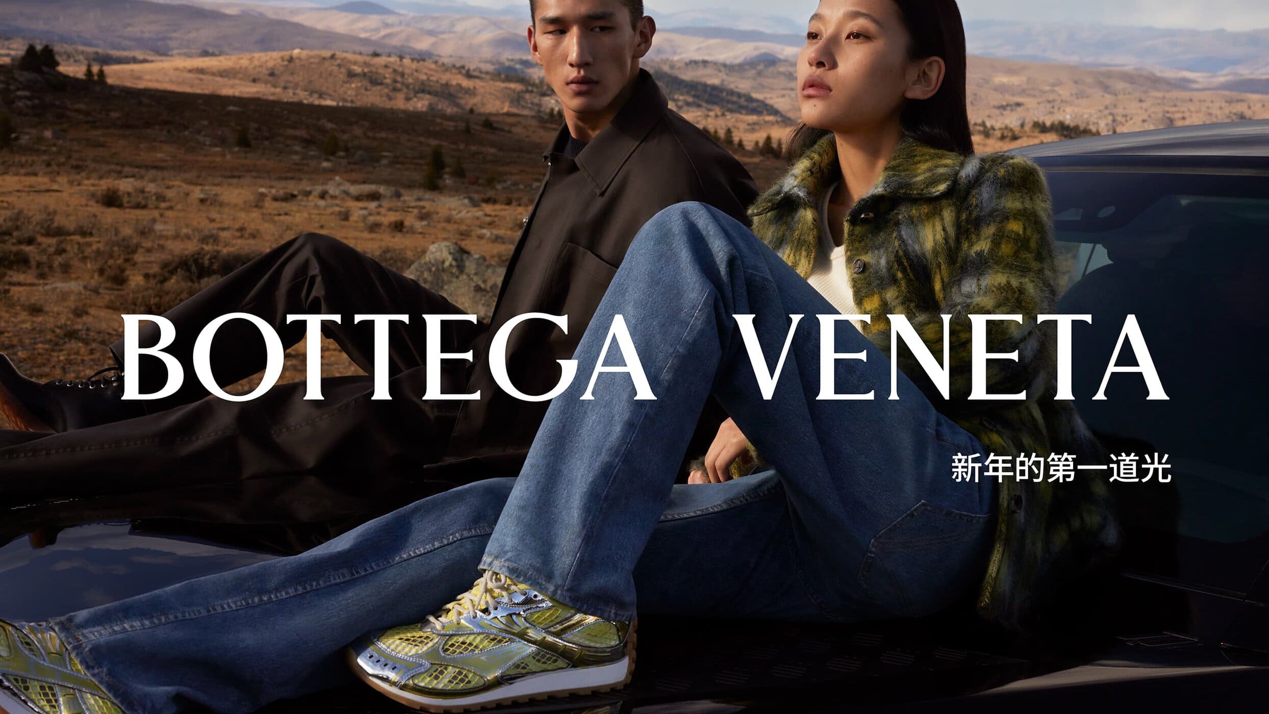 Two adult models pose on a black car in a mountainous, sunlit landscape, showcasing fashion and style with 'Bottega Veneta' text overlay.