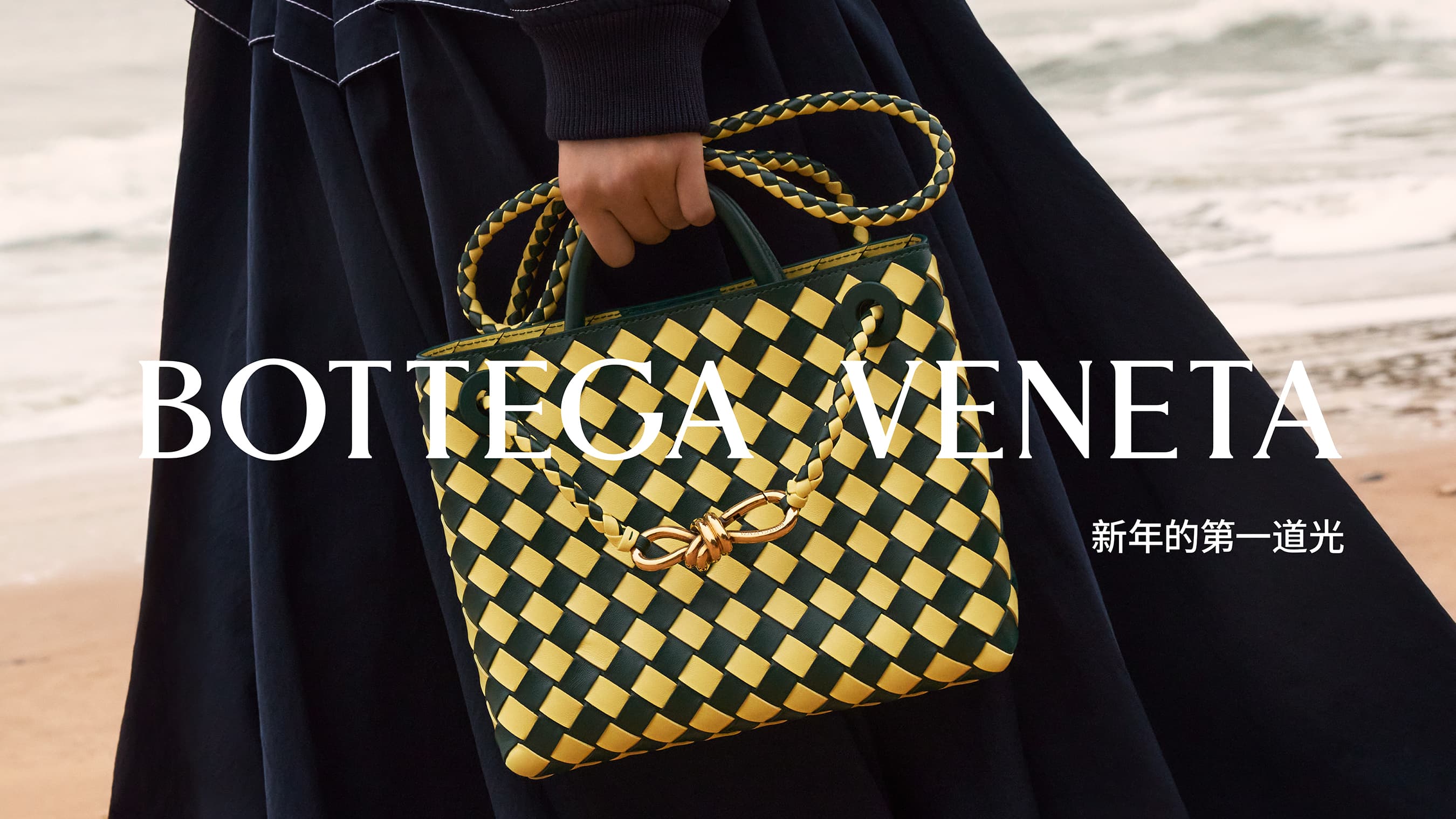 Bottega Veneta woven leather handbag in green and yellow is held in front of a blurred beach scene, with visible brand text.