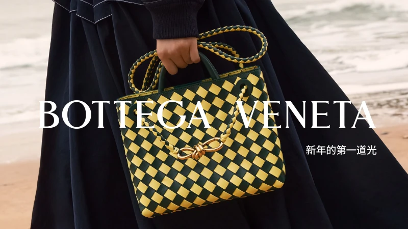 Bottega Veneta woven leather handbag in green and yellow is held in front of a blurred beach scene, with visible brand text.