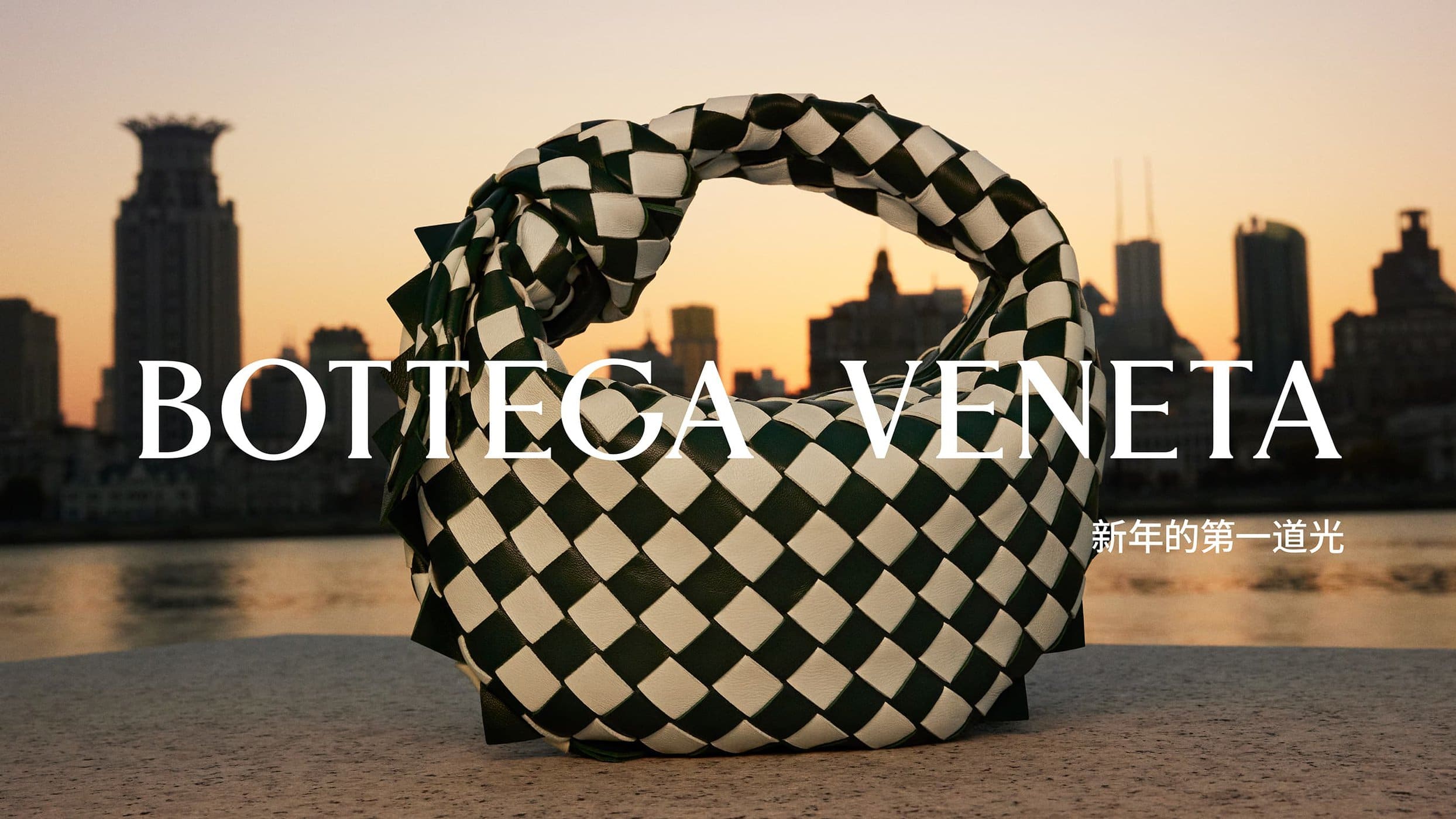 Bottega Veneta handbag in a checkered green and white pattern is displayed against a city skyline and sunset over a body of water.