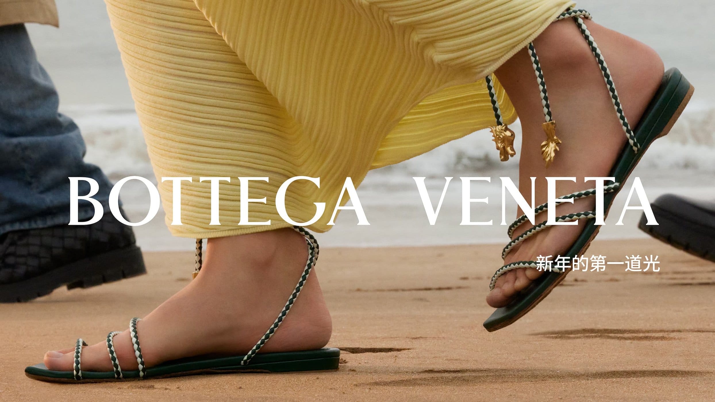 Close-up fashion shot of a woman on a sandy beach wearing green strappy sandals with gold leaf charms, a yellow pleated dress, ocean backdrop.