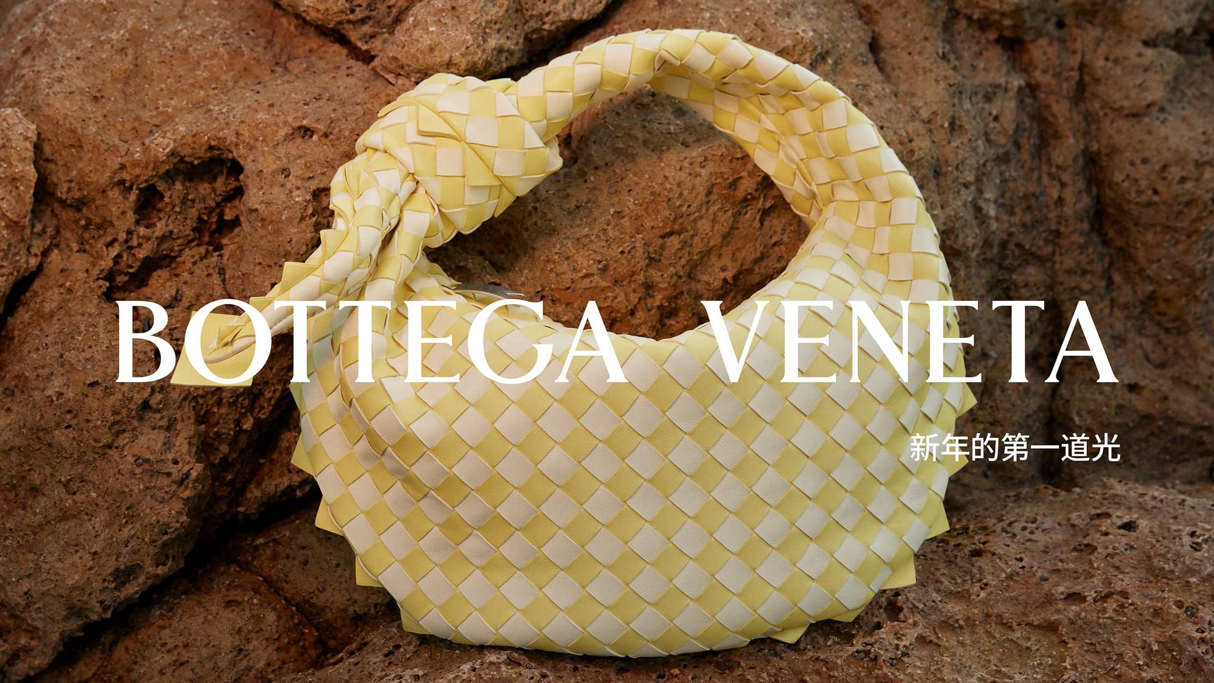 Bottega Veneta woven handbag in light yellow and cream sits against a textured rock background in a fashion product shot.