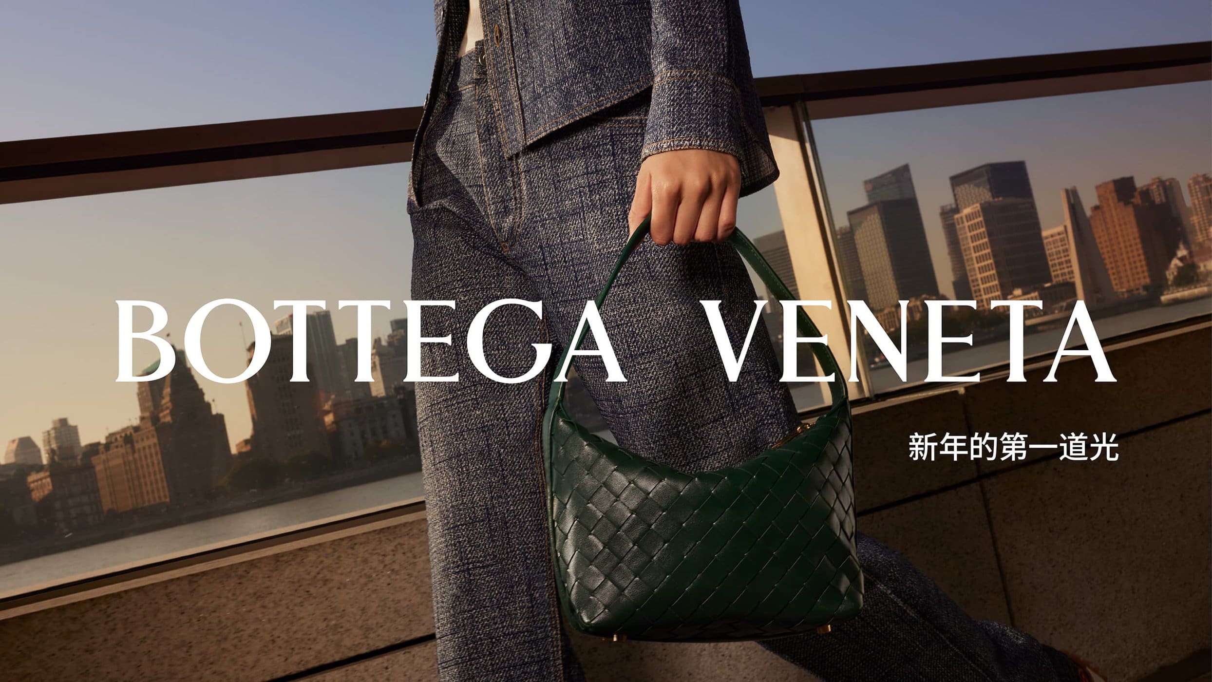 Close-up of a person wearing denim clothing holding a dark green woven leather handbag against a city skyline at sunset.
