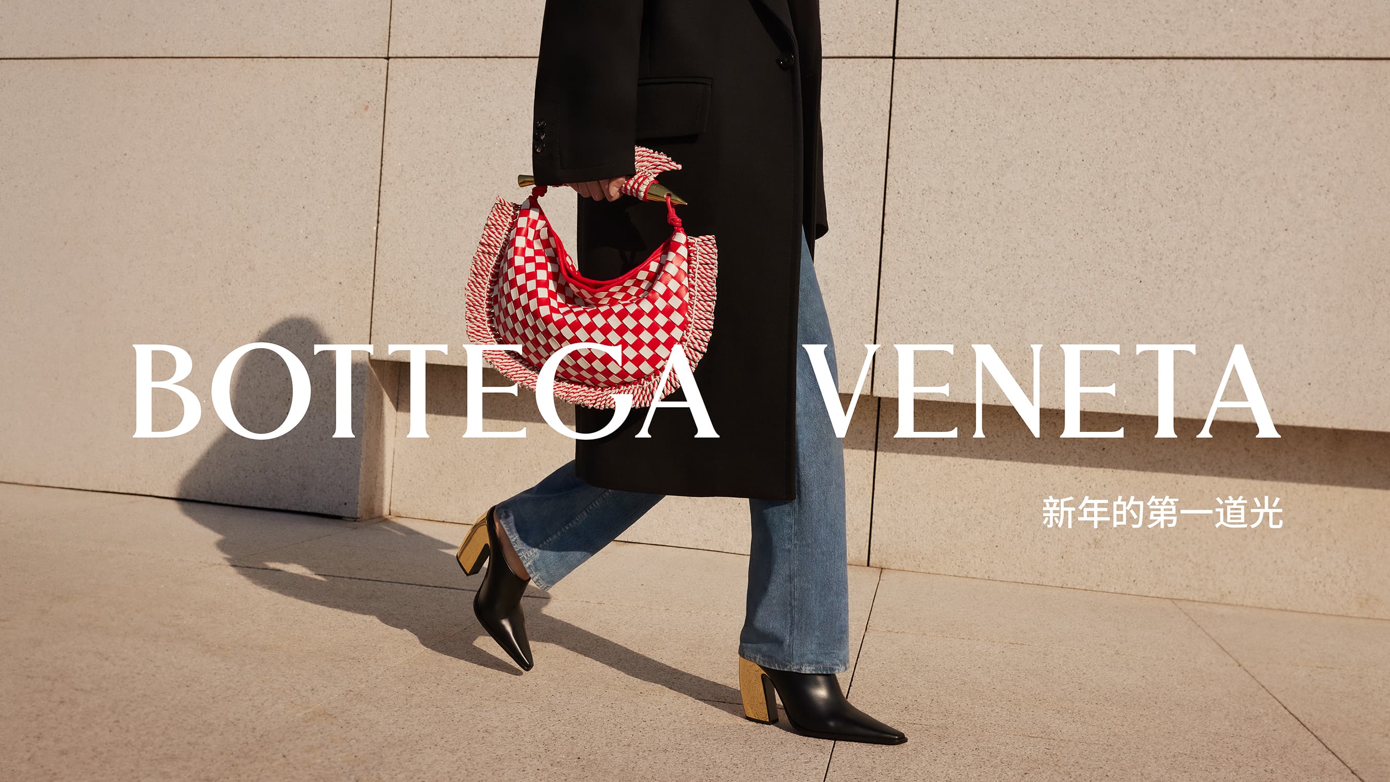 Woman in a black coat walks past a beige wall with a red and white Bottega Veneta bag and black heeled boots, lit by bright sunshine.