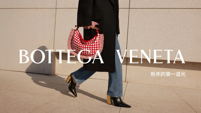 Woman in a black coat walks past a beige wall with a red and white Bottega Veneta bag and black heeled boots, lit by bright sunshine.
