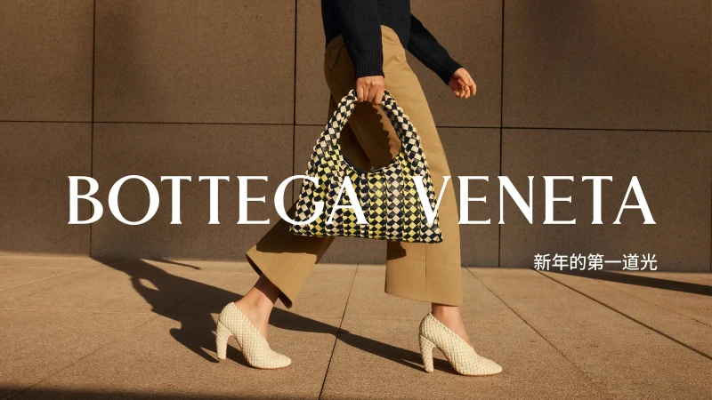 A person walks across a paved outdoor space carrying a woven black, white, and yellow patterned shoulder bag by Bottega Veneta.