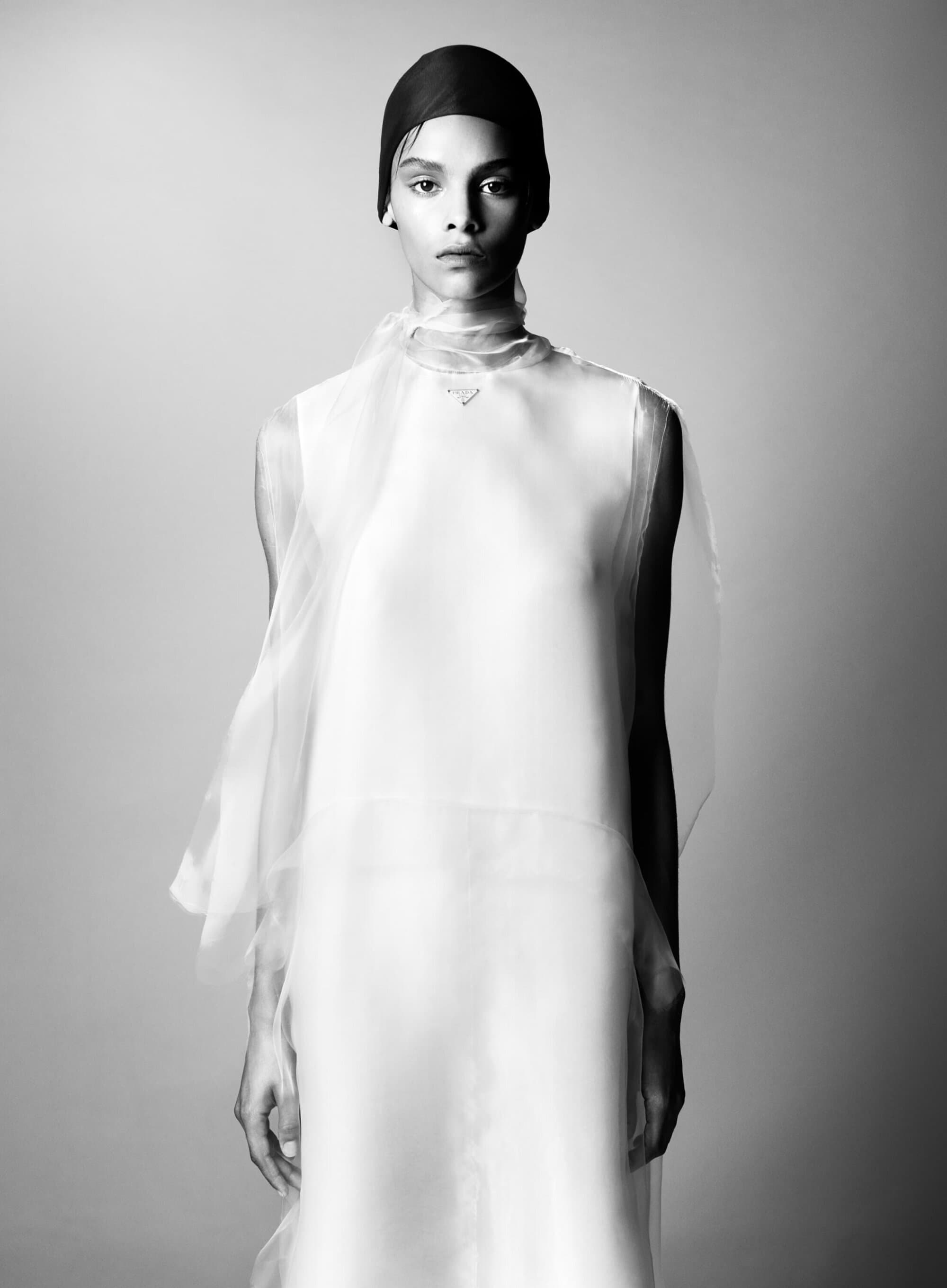 Monochrome portrait of an androgynous person with a dark head covering, wearing a white dress against a neutral backdrop.