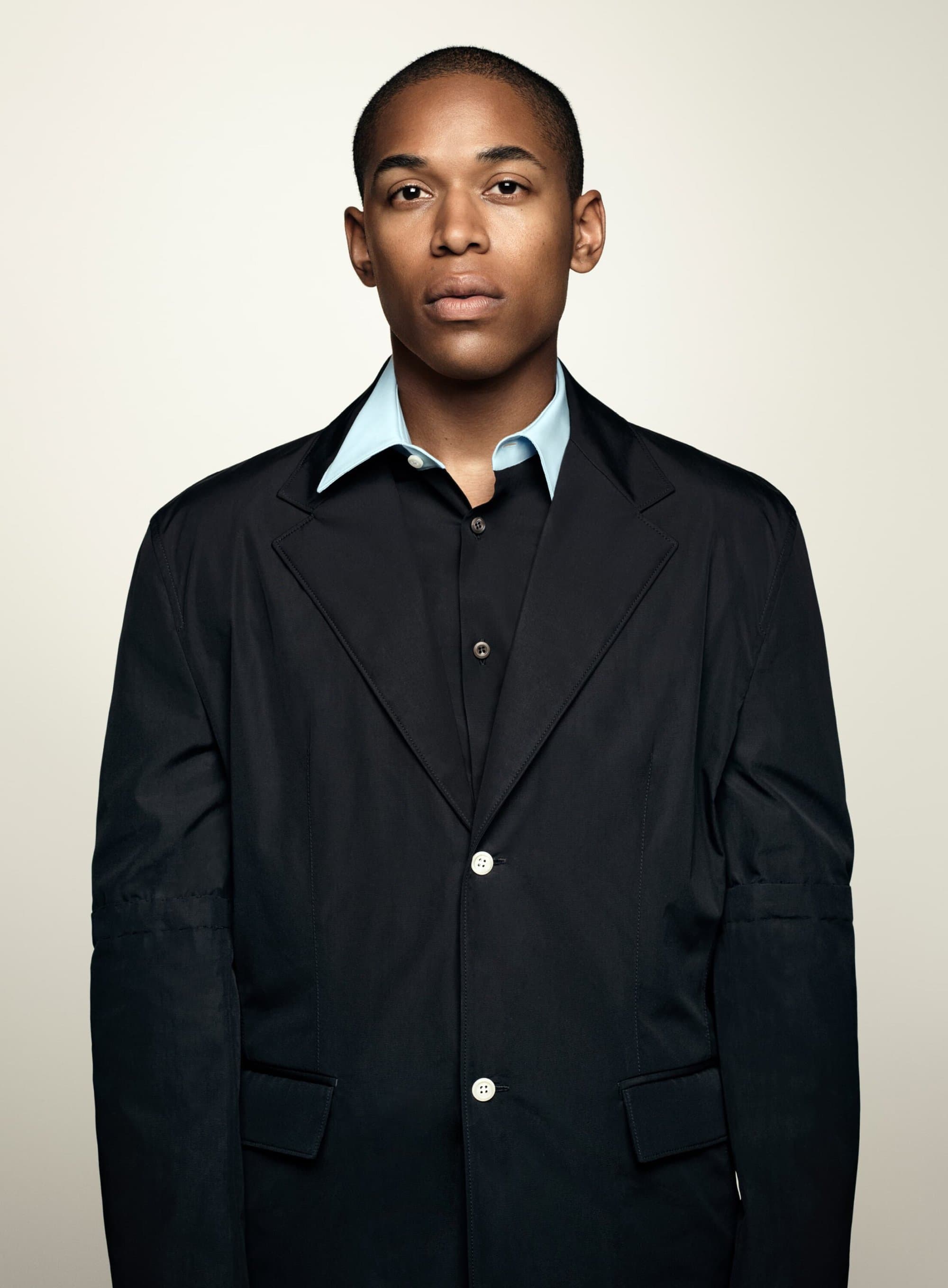 Portrait of an adult man wearing a black blazer and light blue collared shirt against a neutral beige backdrop in a studio setting.
