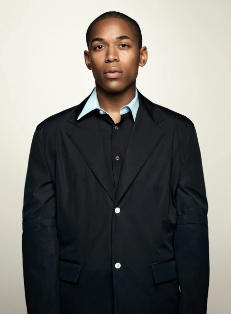 Portrait of an adult man wearing a black blazer and light blue collared shirt against a neutral beige backdrop in a studio setting.