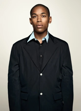 Portrait of an adult man wearing a black blazer and light blue collared shirt against a neutral beige backdrop in a studio setting.