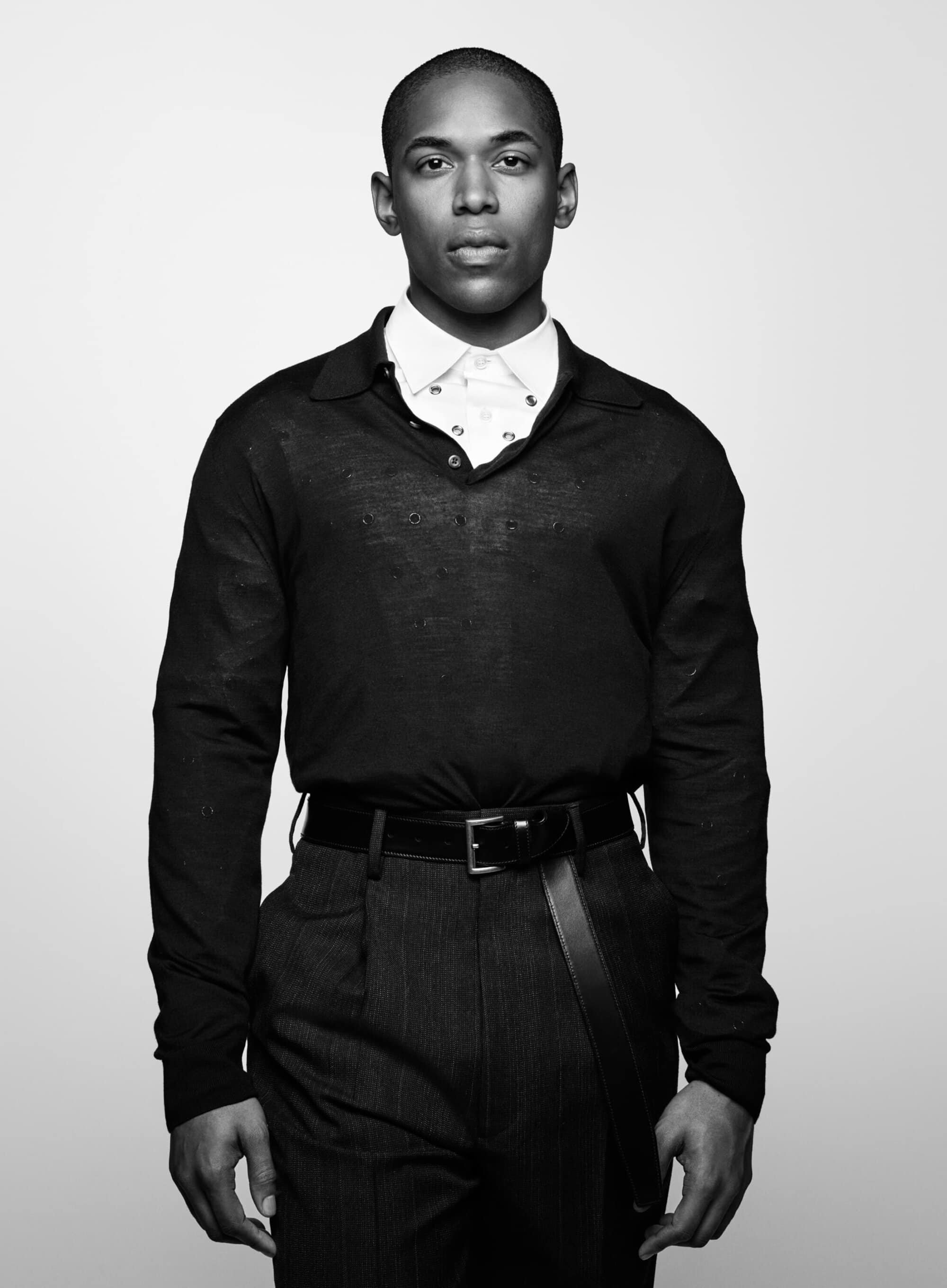 Portrait of a Black man in a white button-down shirt and dark sweater, wearing pinstripe pants and a black belt against a white background.