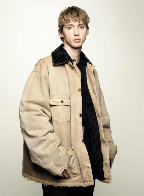 Young man in a tan workwear jacket with a dark collar, captured in a medium shot against a bright, neutral background in a fashion photography style.