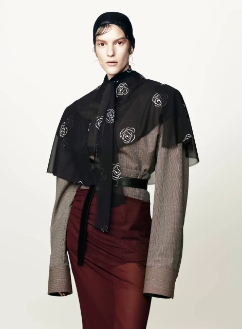Fashion shot of a model wearing a brown plaid top, black cape with white floral design, maroon skirt, and black head covering.