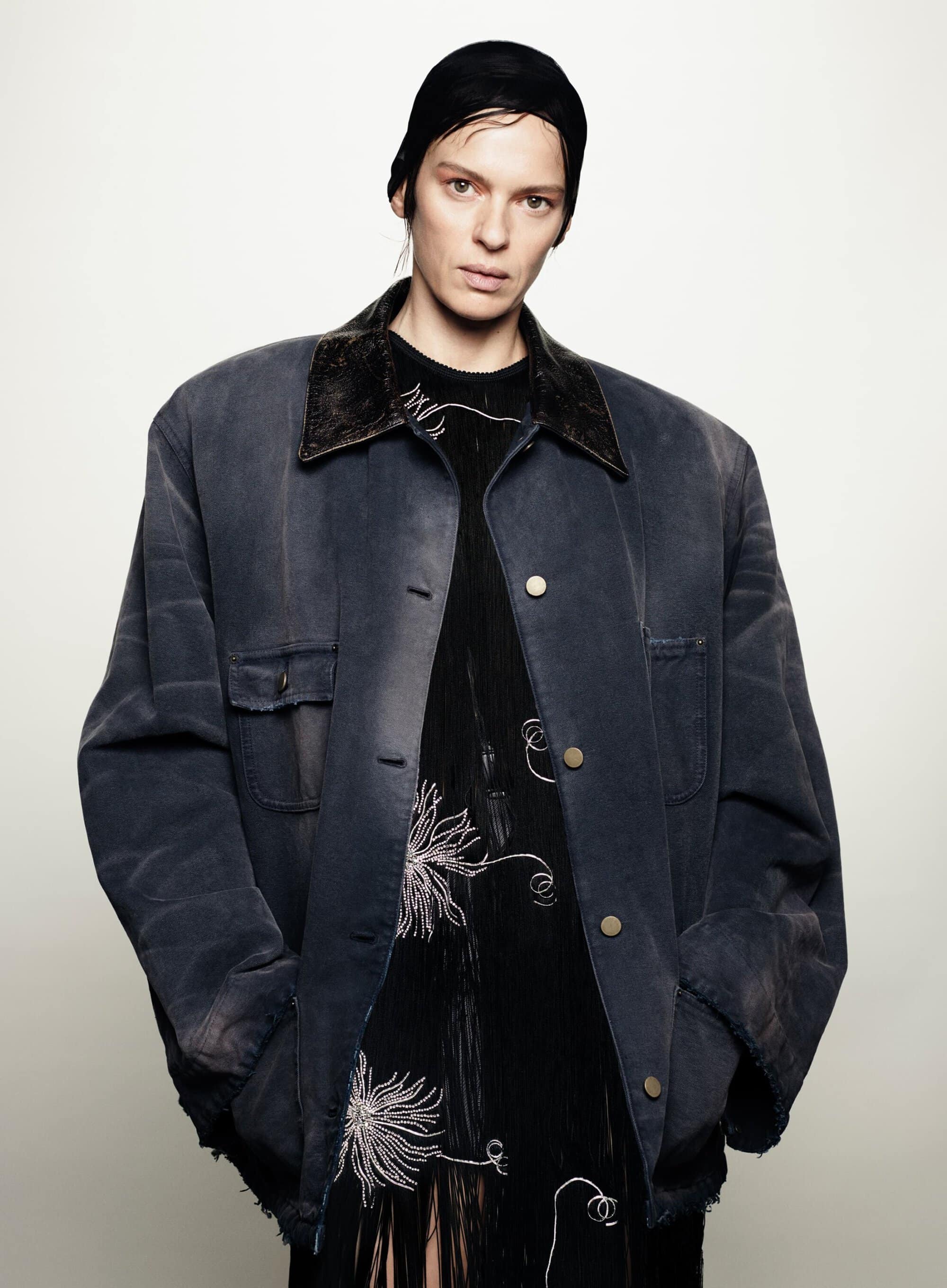 Studio portrait of a woman wearing a black head covering, oversized jacket, and black dress with white floral embroidery against a white backdrop.