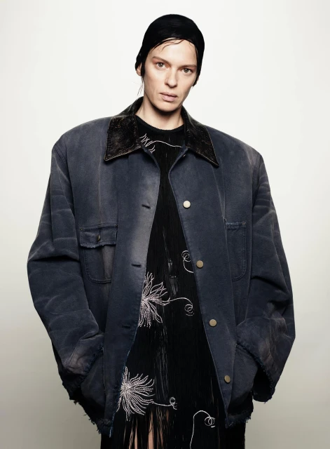 Studio portrait of a woman wearing a black head covering, oversized jacket, and black dress with white floral embroidery against a white backdrop.