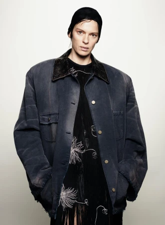 Studio portrait of a woman wearing a black head covering, oversized jacket, and black dress with white floral embroidery against a white backdrop.