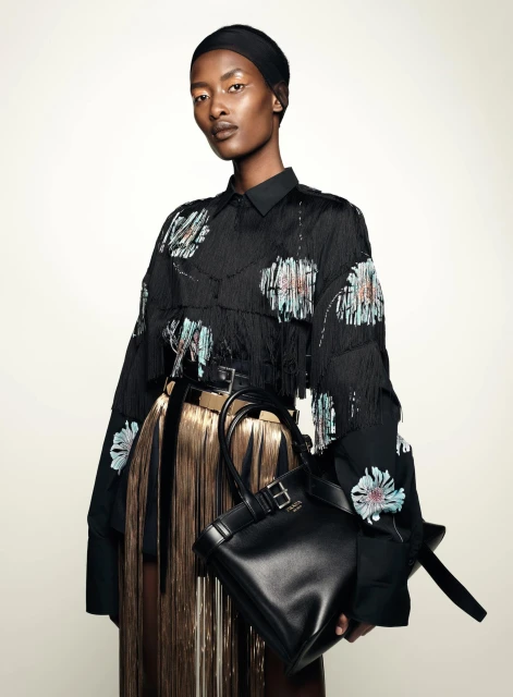 Fashion editorial shot of a Black female model in a black floral fringe shirt, gold fringe skirt, black belt and black leather handbag.