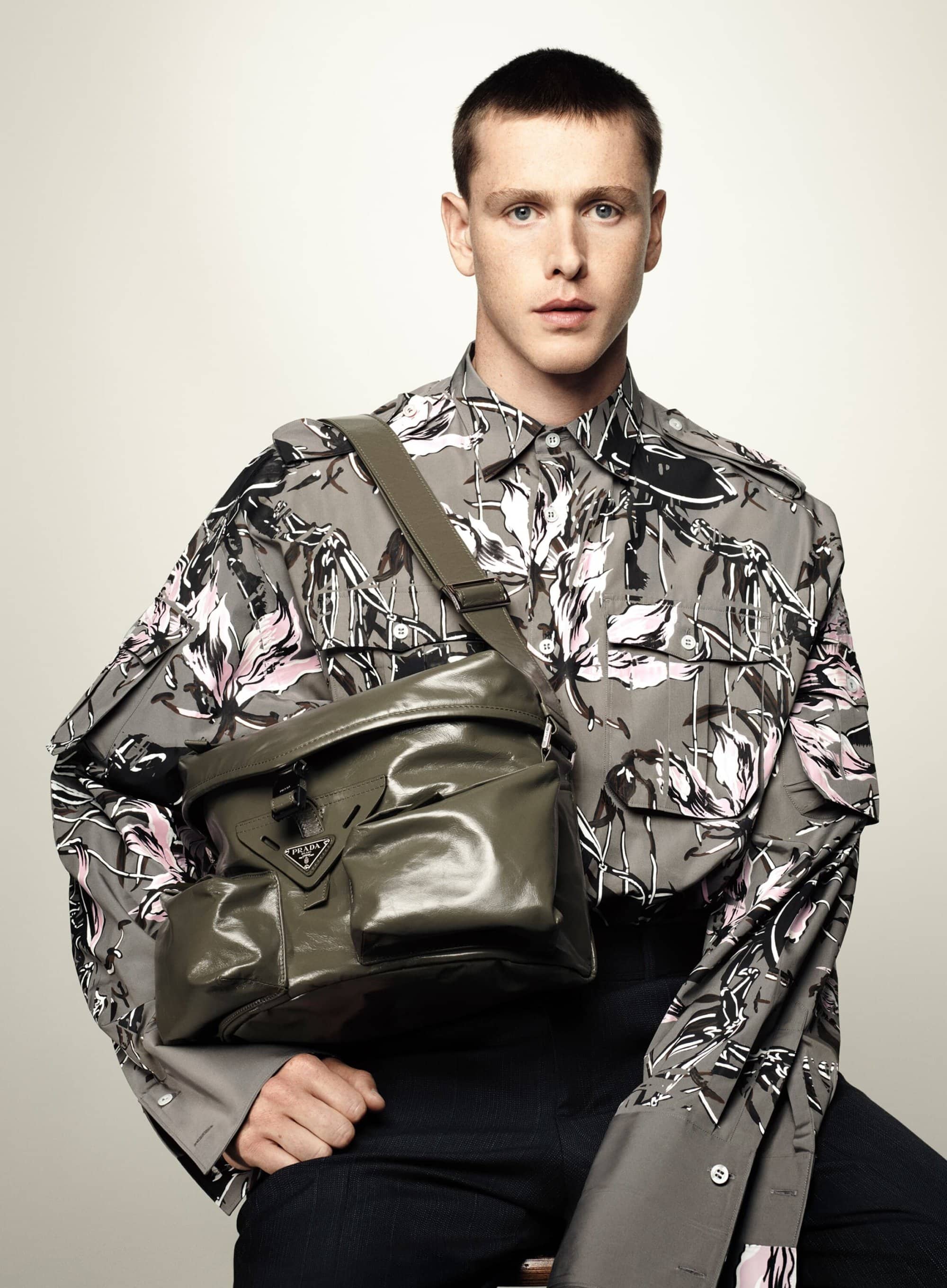 Man in floral shirt poses with an olive-green Prada bag slung across his body in a studio fashion shot against a neutral backdrop.