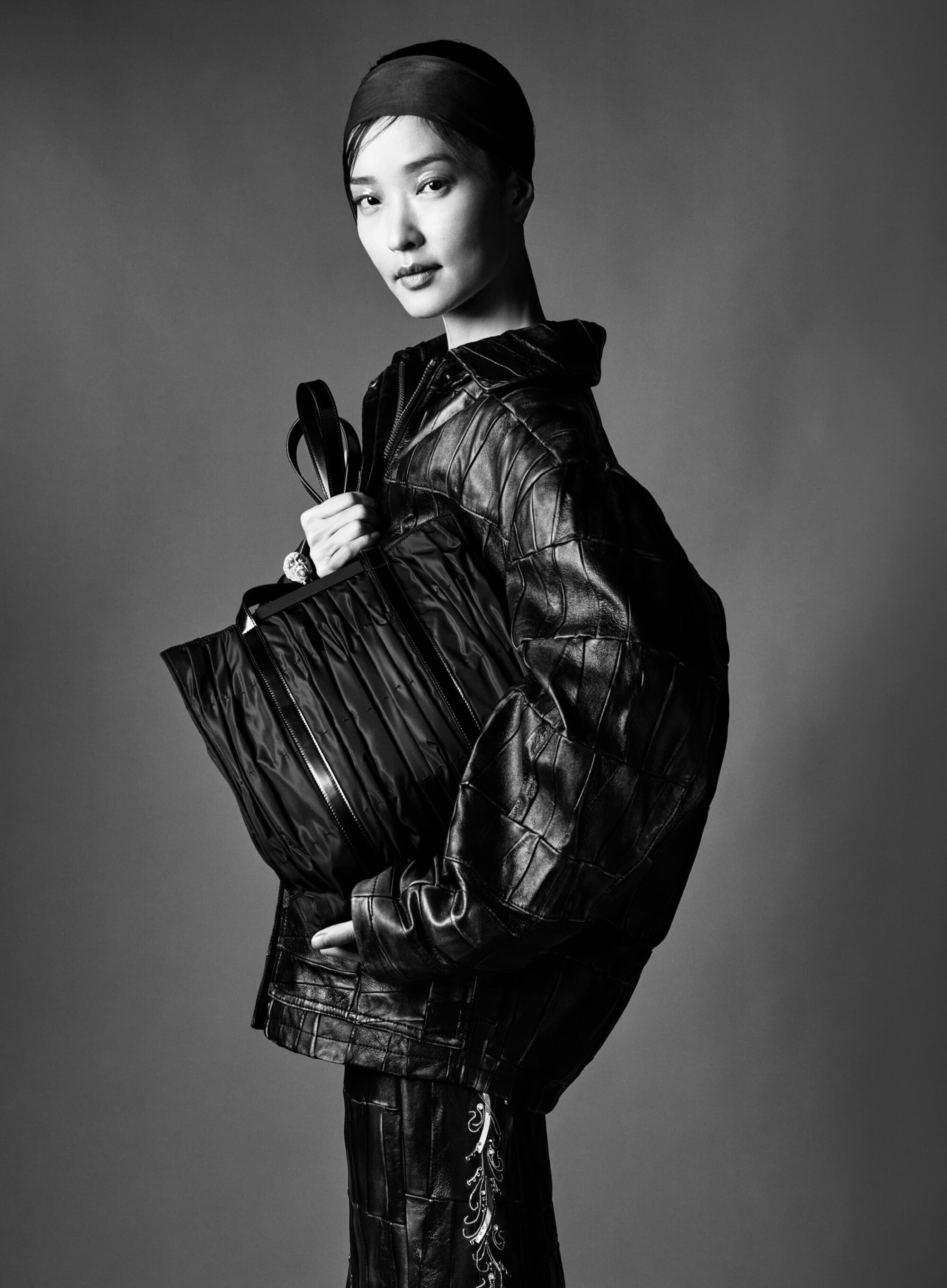 Black and white portrait of an Asian woman with short hair, wearing a leather jacket and holding a tote bag, against a grey background.