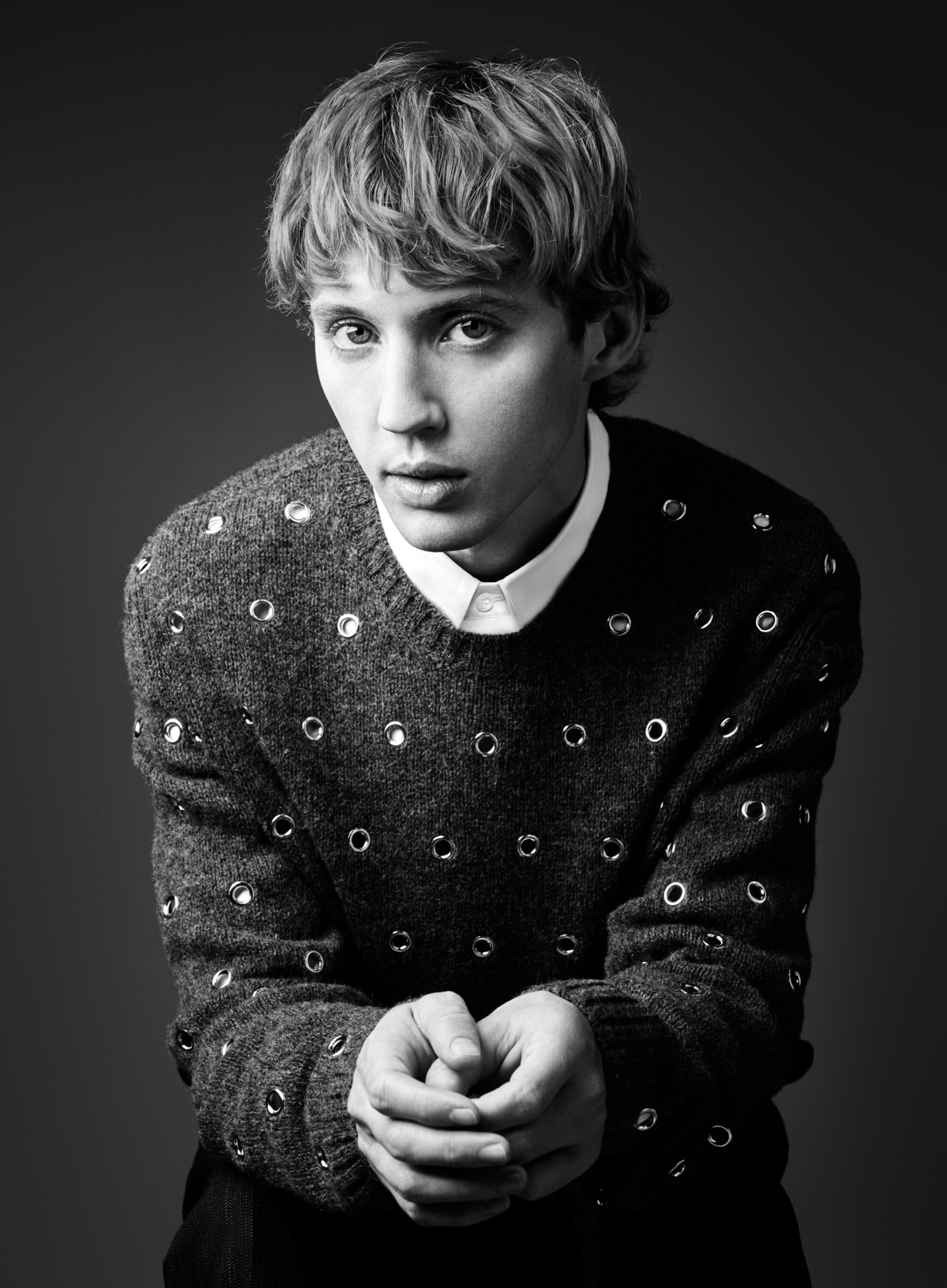Black and white portrait of a young adult male with wavy blonde hair, wearing a patterned sweater and collared shirt. Indoors, medium shot.