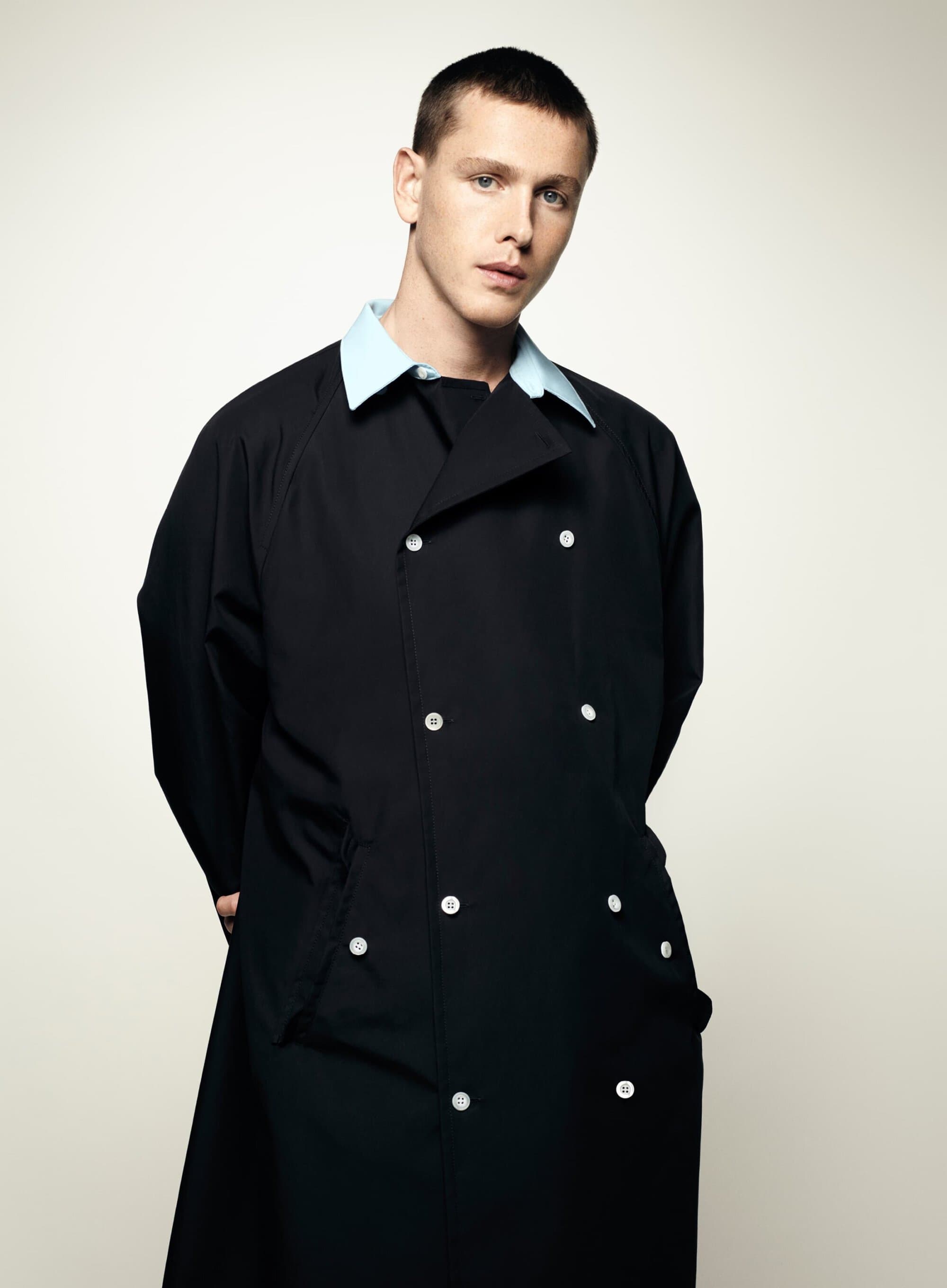 A young, fair-skinned male poses in a photo studio. He is wearing a long, double-breasted black coat with light blue collar.