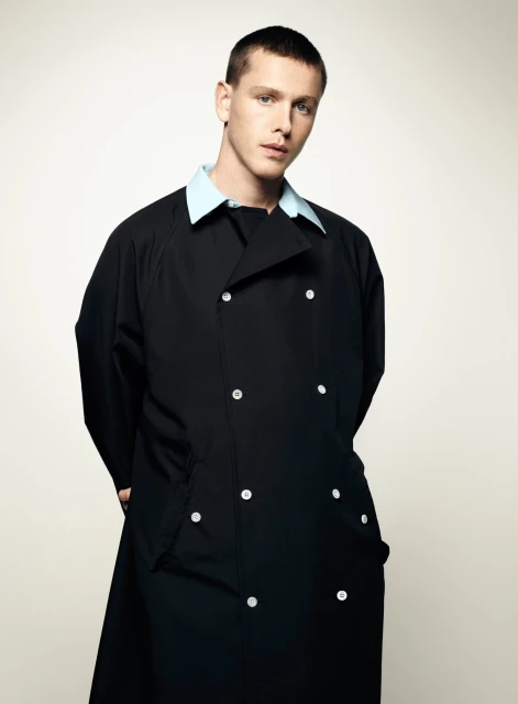 A young, fair-skinned male poses in a photo studio. He is wearing a long, double-breasted black coat with light blue collar.