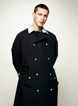 A young, fair-skinned male poses in a photo studio. He is wearing a long, double-breasted black coat with light blue collar.