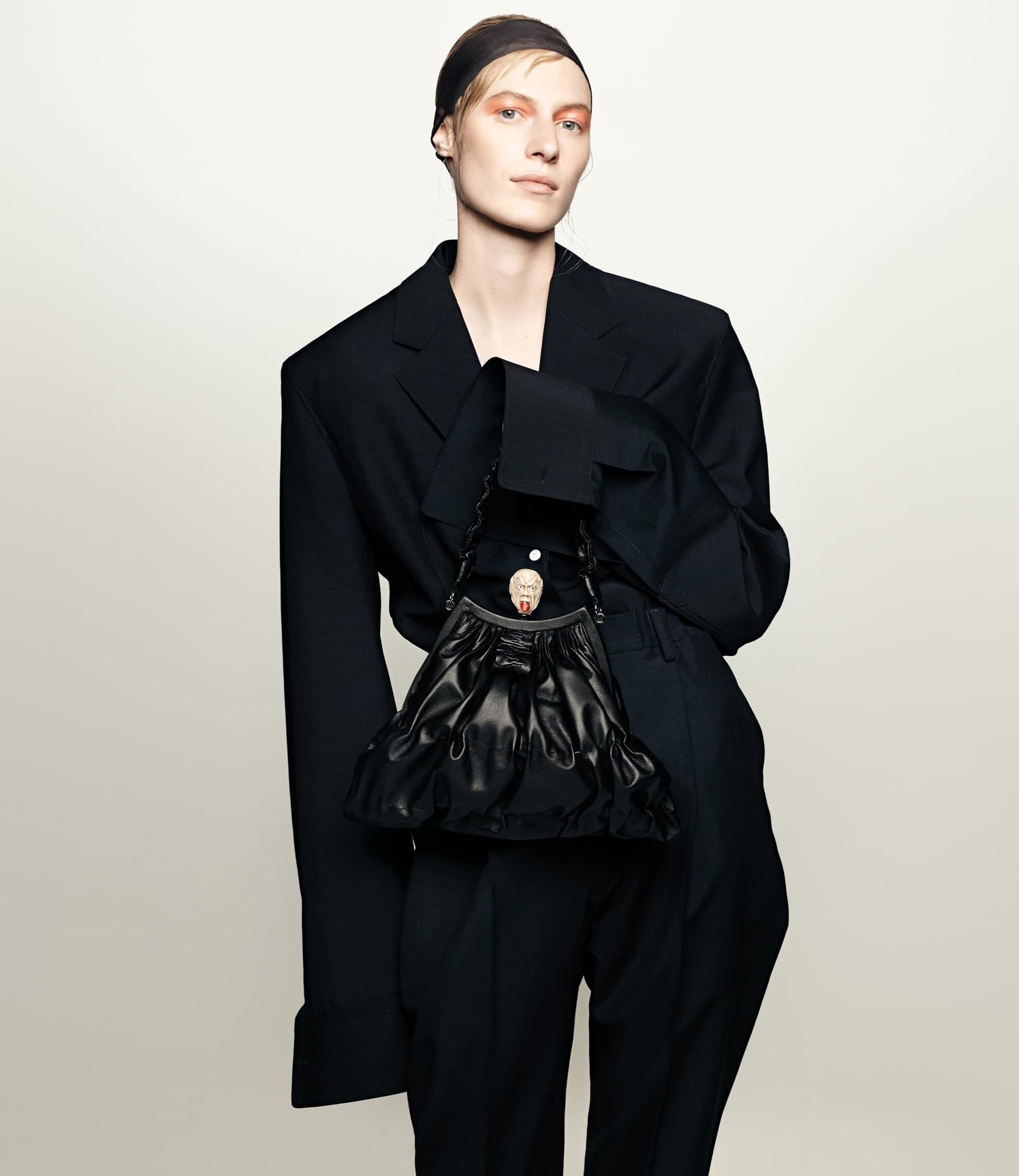 Fashion portrait of androgynous person in a black oversized suit with a black headscarf holding a black leather handbag in studio.
