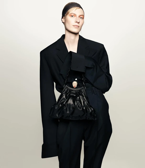 Fashion portrait of androgynous person in a black oversized suit with a black headscarf holding a black leather handbag in studio.