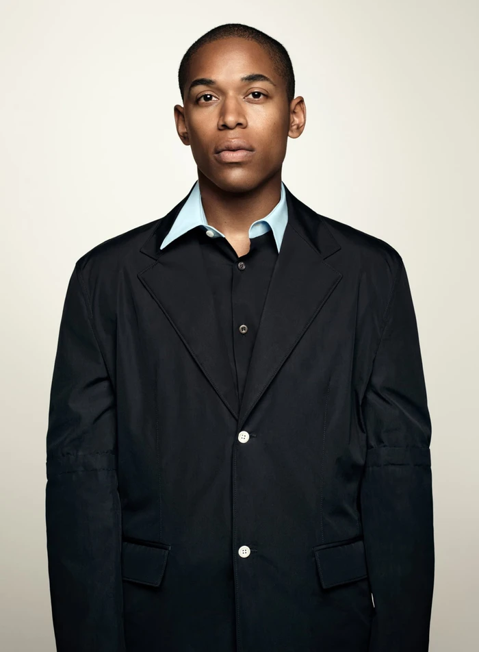Portrait of a man in a black jacket and blue shirt against a plain neutral background for Prada Spring 2024 campaign