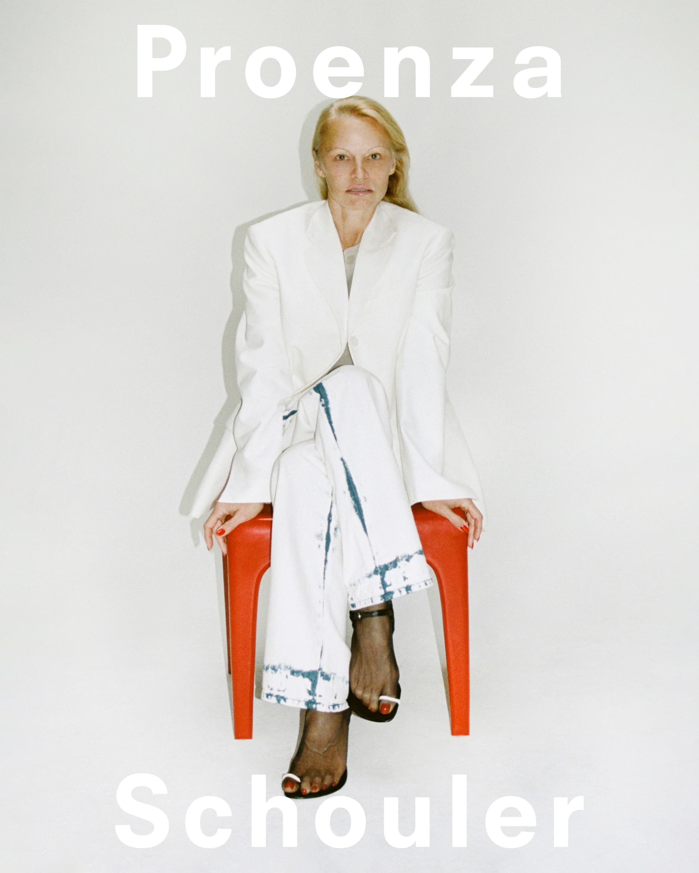 Woman in white suit sits on a red stool in a studio with the words Proenza Schouler in white at the top and bottom.