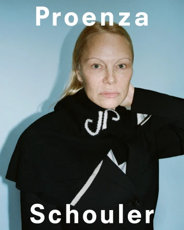 Close-up of a blonde woman wearing a black coat with white detailing against a light blue background. Fashion brand logo visible.