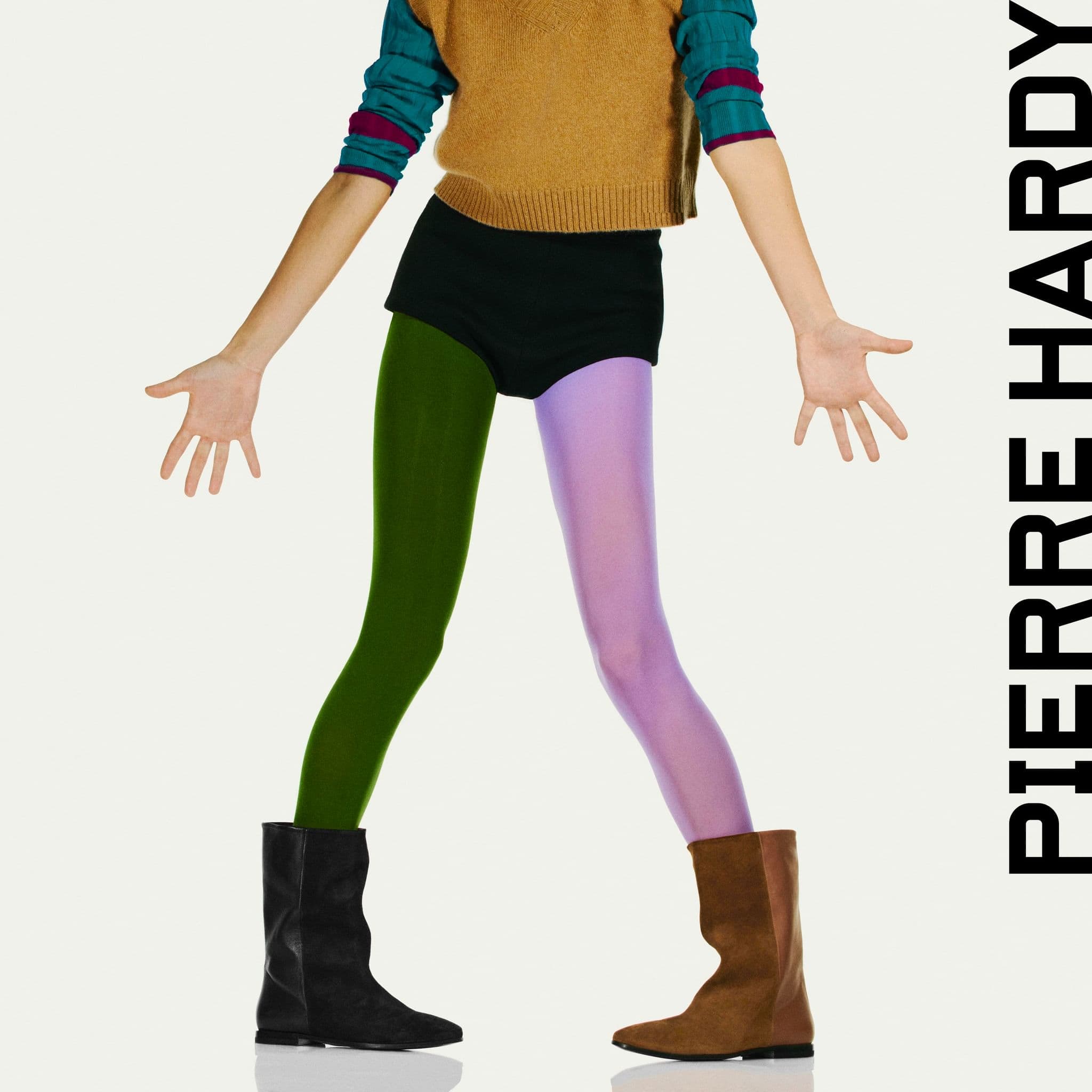 Fashion shot of androgynous model in sweater, black shorts, different colored tights, and ankle boots standing on white seamless.