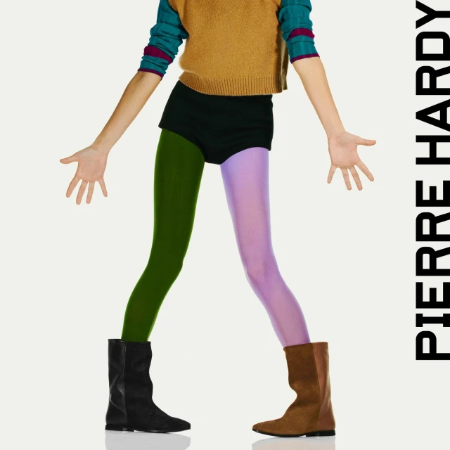 Fashion shot of androgynous model in sweater, black shorts, different colored tights, and ankle boots standing on white seamless.