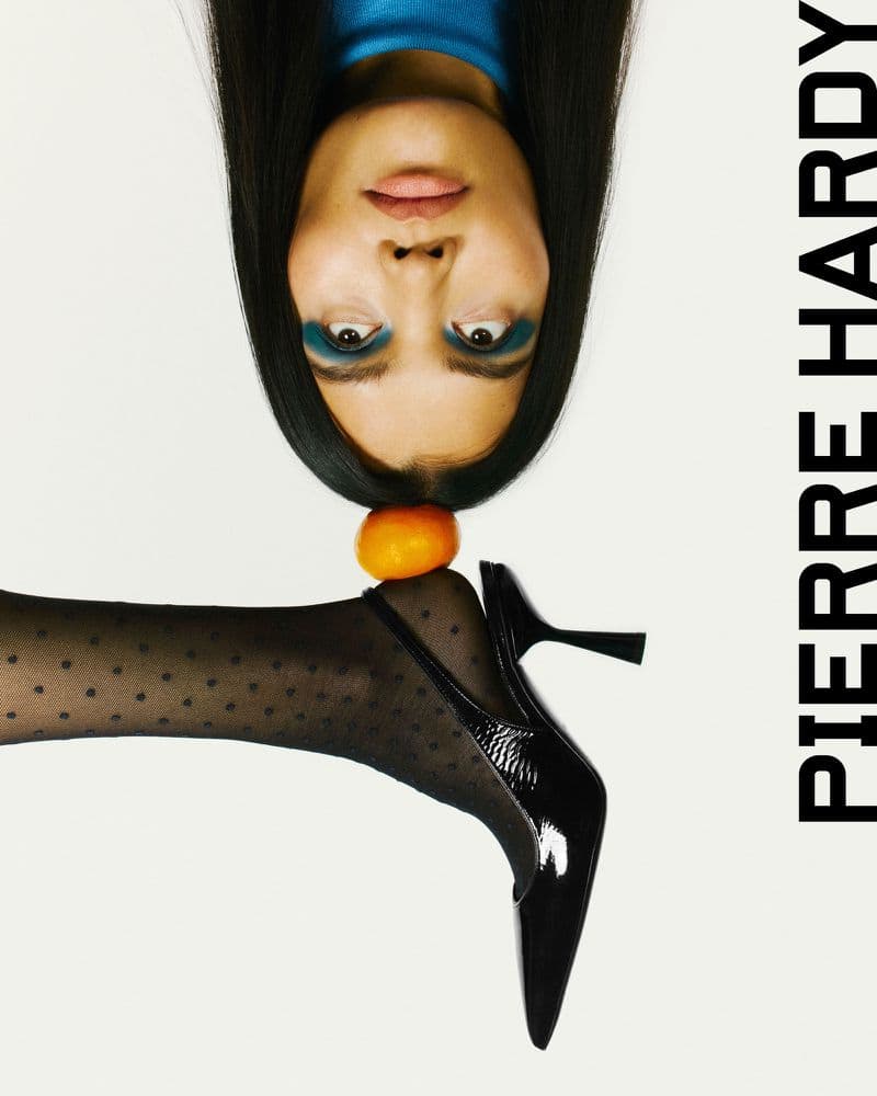 A woman poses with a tangerine balanced on her foot wearing a black high heel and patterned stockings, against a white background.