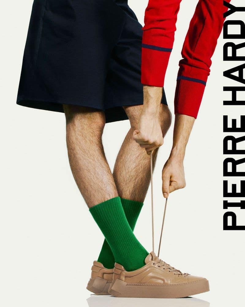 Close-up studio shot of a man tying the shoelaces of a tan sneaker, wearing dark shorts and green socks, with a partial view of a red shirt.